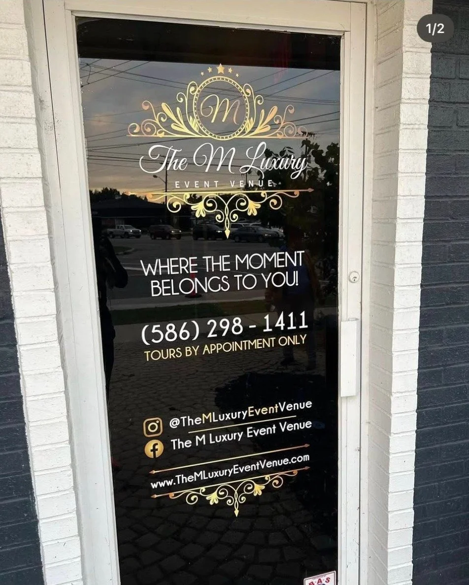 Glass door with the logo and contact information of The M Luxury Event Venue. The logo features a gold decorative crest with a central letter 'M'. The text includes the venue's slogan, phone number, social media handles, and website, with reflections