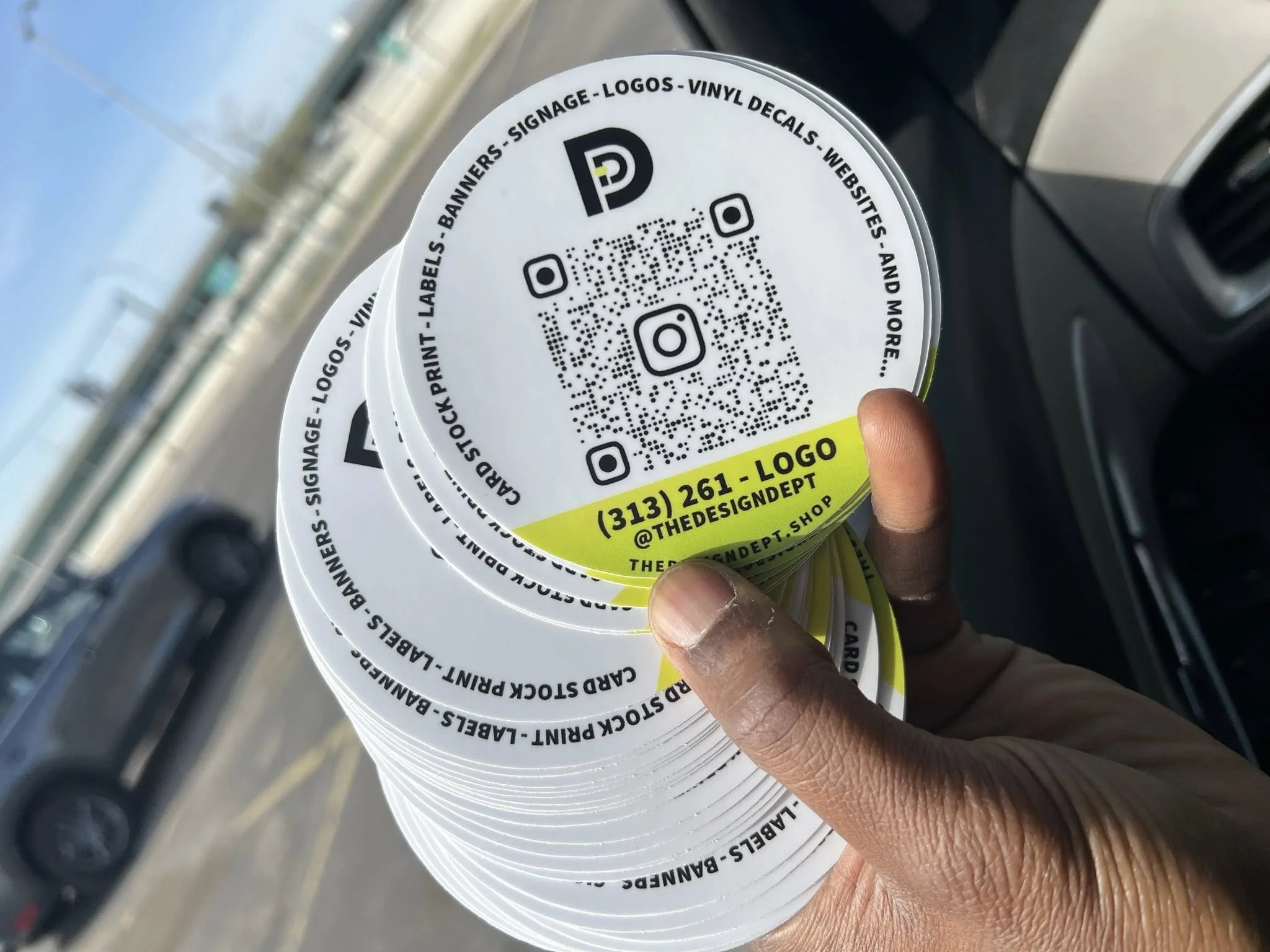Hand holding a stack of round promotional cards with black, white, and yellow design, featuring QR codes, social media icons, and contact information, inside a vehicle.
