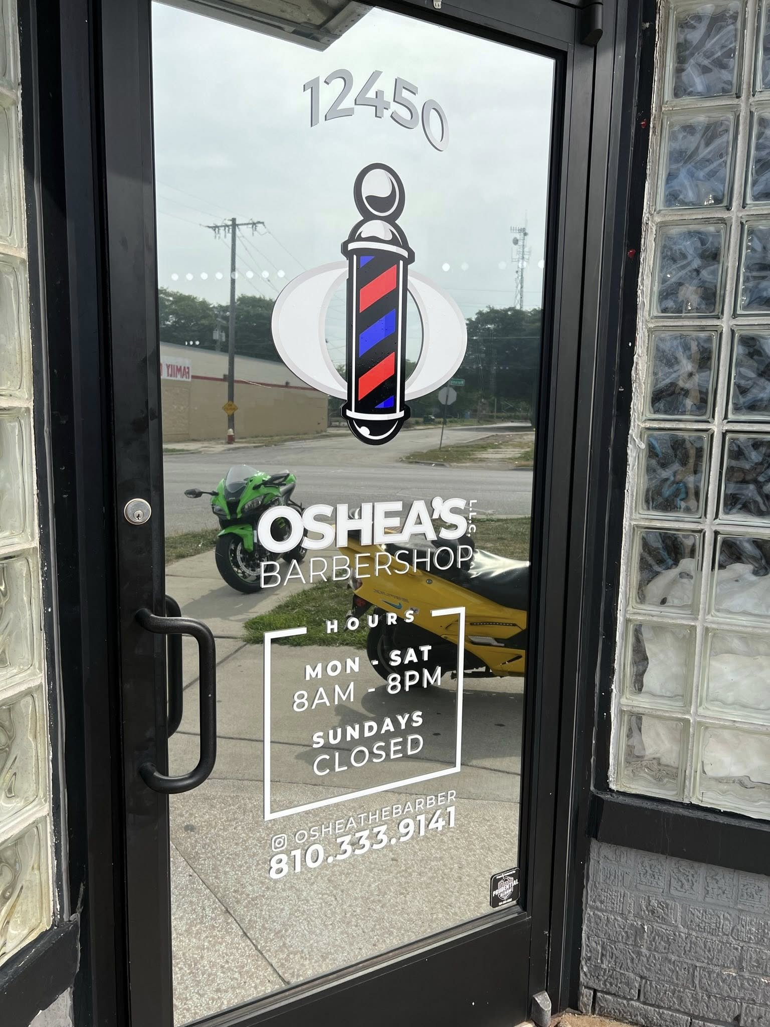 Glass door with barber shop hours, logo, and contact information, and reflections of street, motorcycle, and yellow vehicle outside.