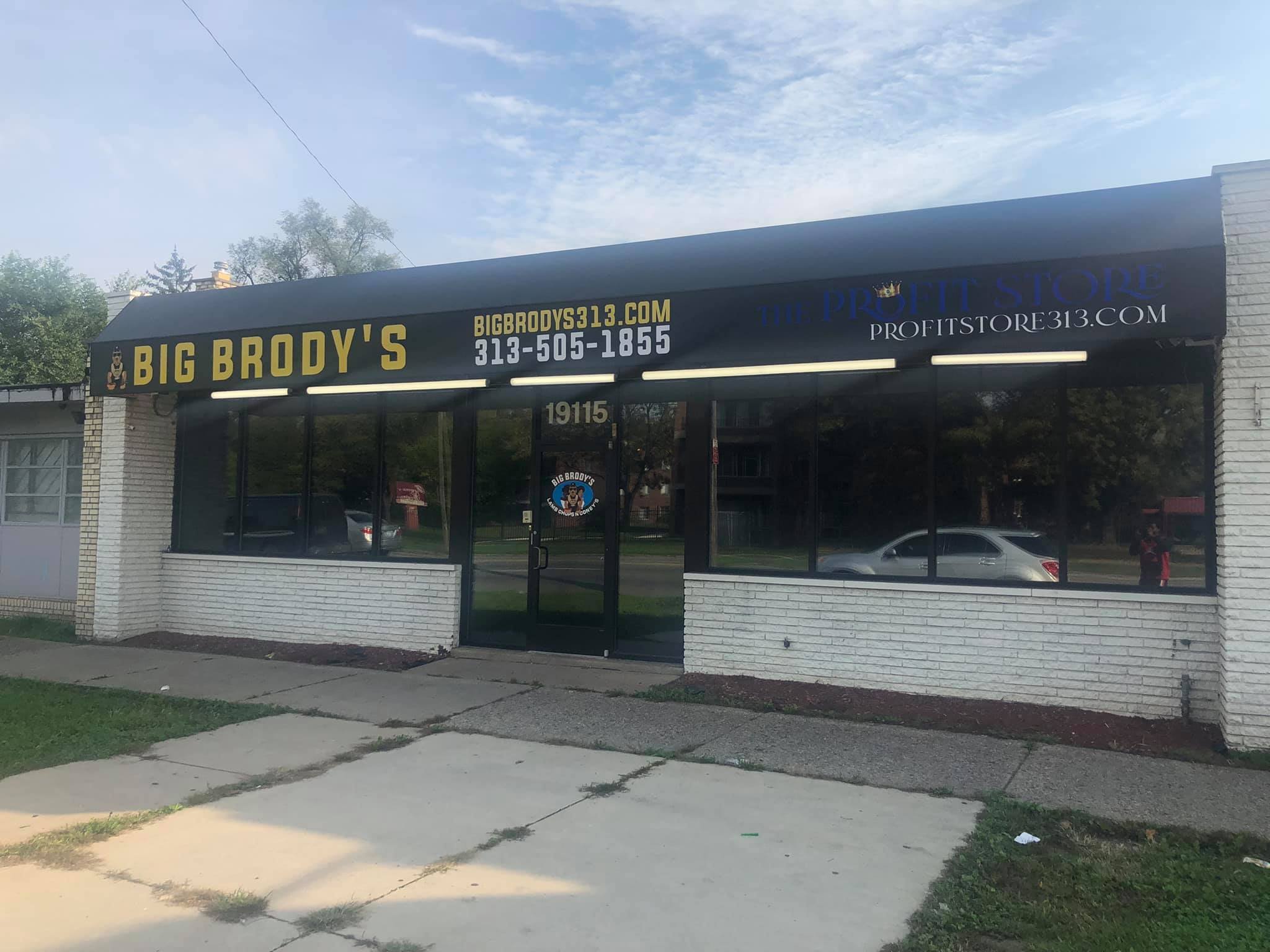 Front view of a brick storefront with large windows and a black awning displaying yellow and blue text, including the name 'Big Brody's' and website information, with a sidewalk and street reflected in the windows.
