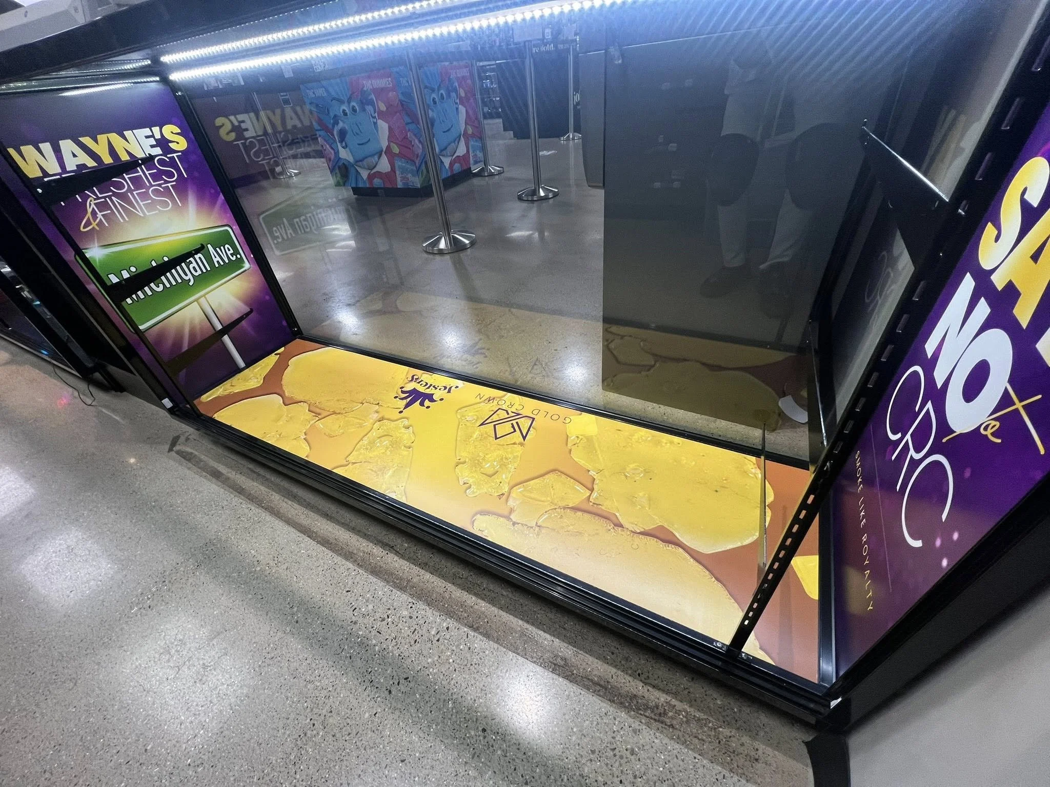 Empty refrigerated display case with glass front, positioned at an airport or shopping mall, with advertisements on the side and roast, yellow, and white marbled cheese blocks inside.