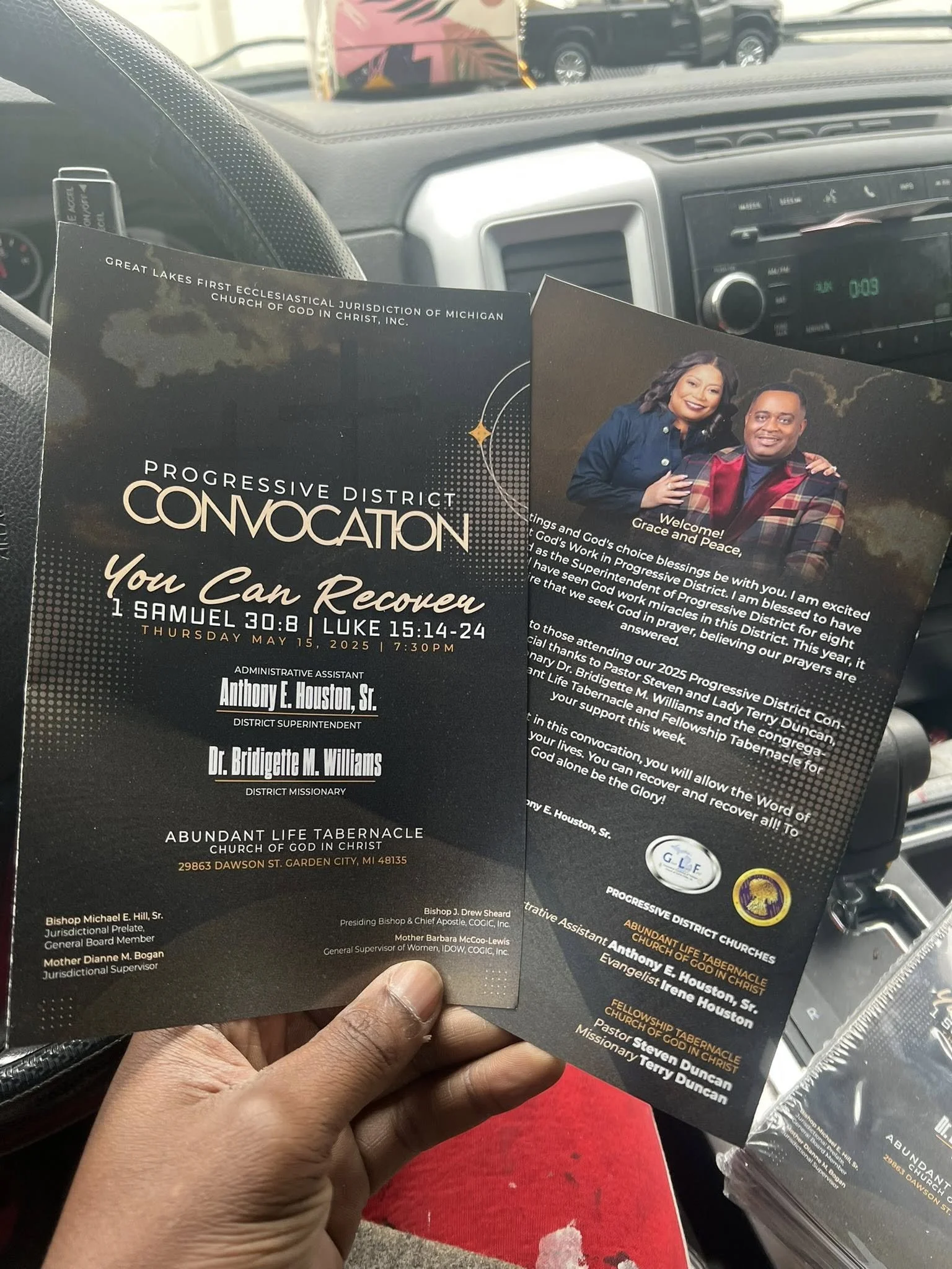 A person holds two event flyers inside a vehicle. One flyer is for a religious event titled "Progressive District Convocation: You Can Recover," held on May 15, 2025. The other flyer features a photo of two people, Grace and Pastor Steven Duncan, wit