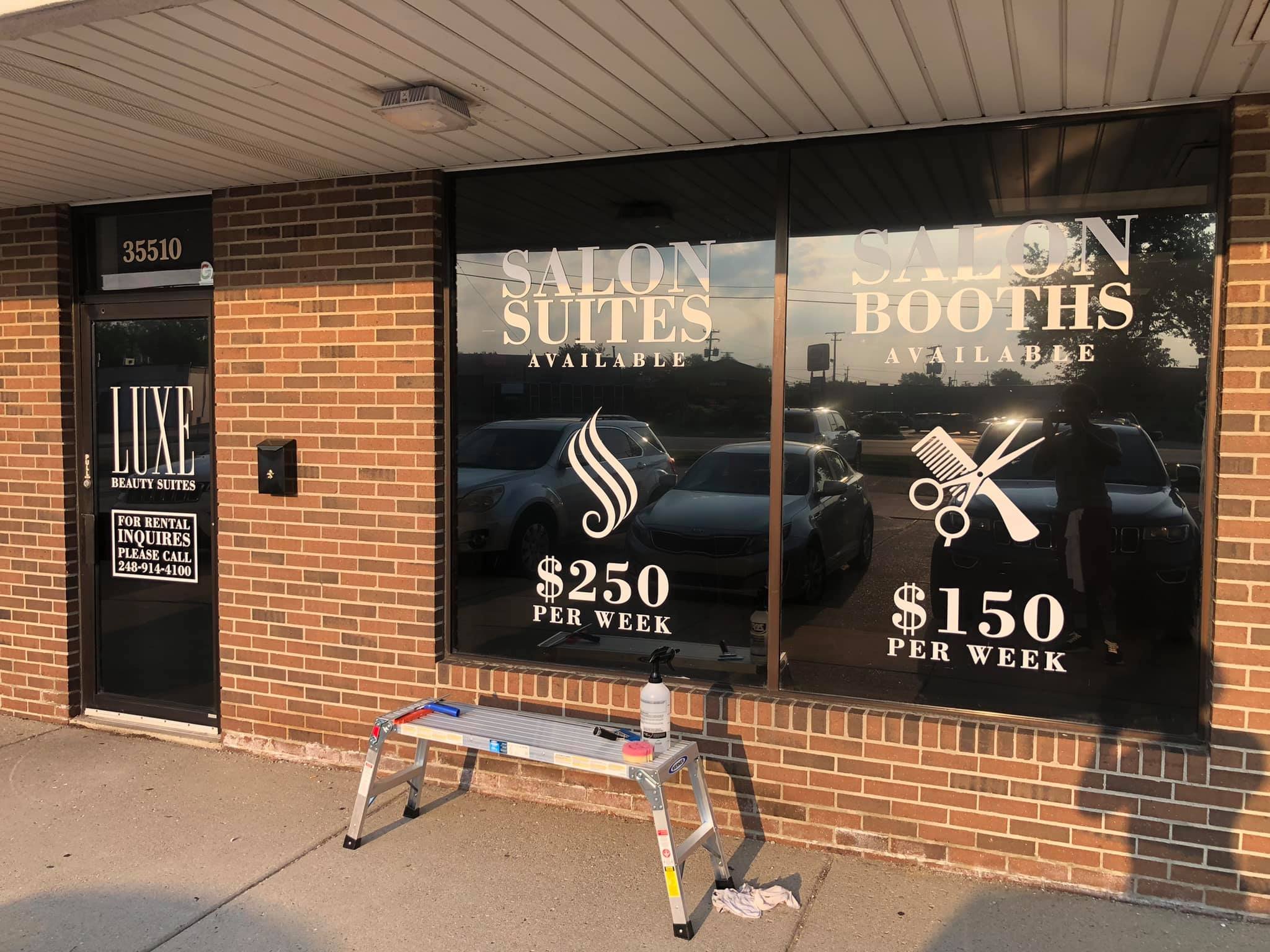 Beauty salon storefront with large windows displaying services and prices, brick exterior, entrance door with sign, a small ladder with cleaning supplies outside.