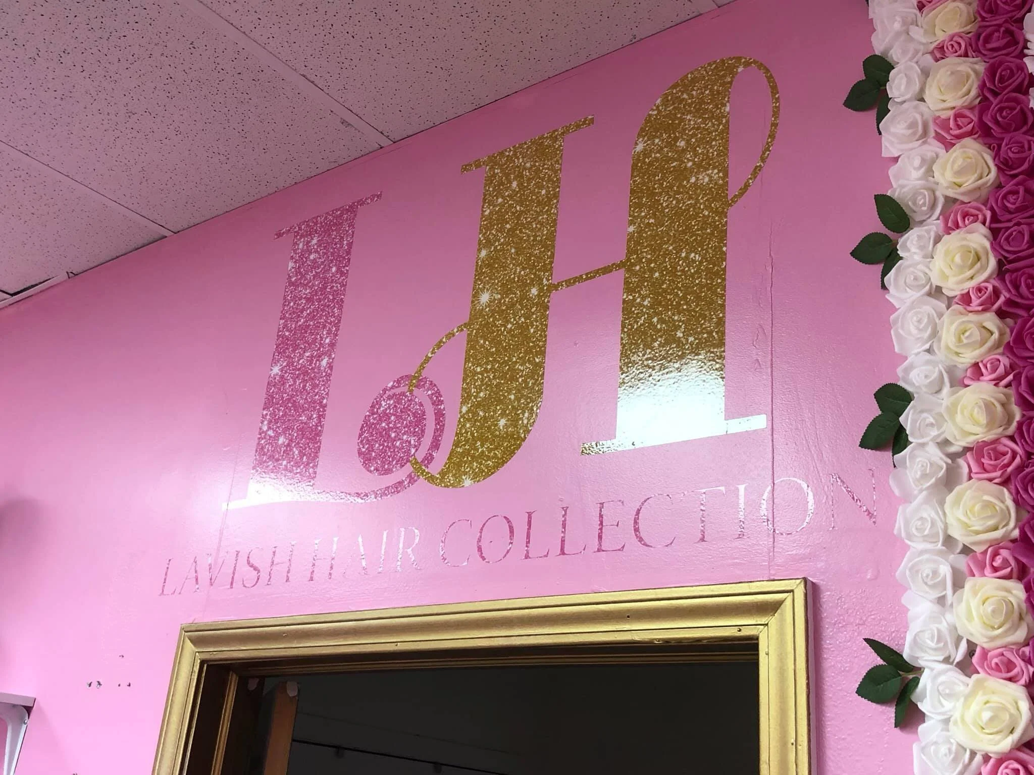 Pink wall with gold and pink glitter letters spelling 'LO' and a floral decoration of white and pink roses to the right.