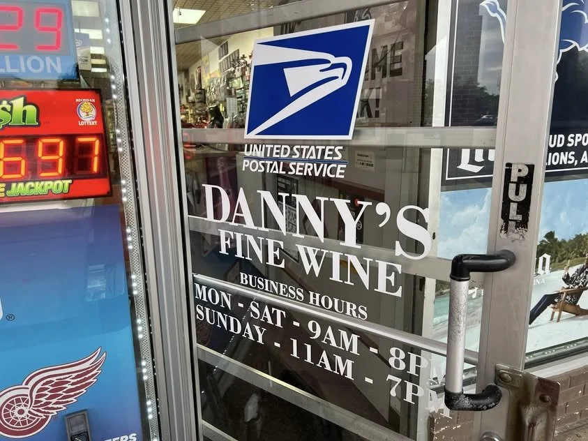 Glass door with signage for Danny's Fine Wine, USPS, and lottery, showing business hours and USPS logo.