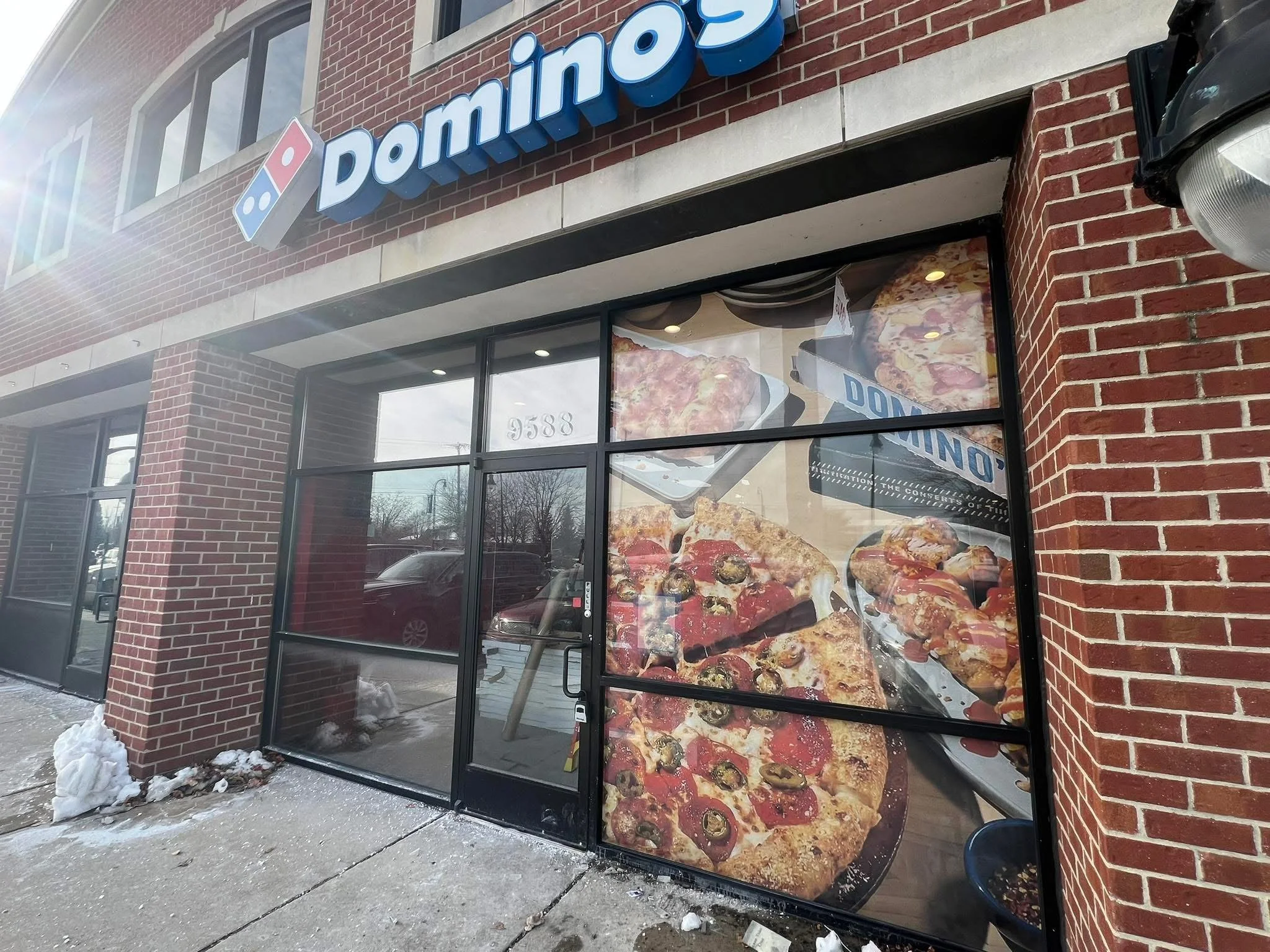 Exterior of a Domino's Pizza storefront with large window display showcasing images of pizza, pizza slices, and garlic bread. The Domino's logo and name are visible above the entrance, which has a glass door. Snow on the ground indicates winter weath