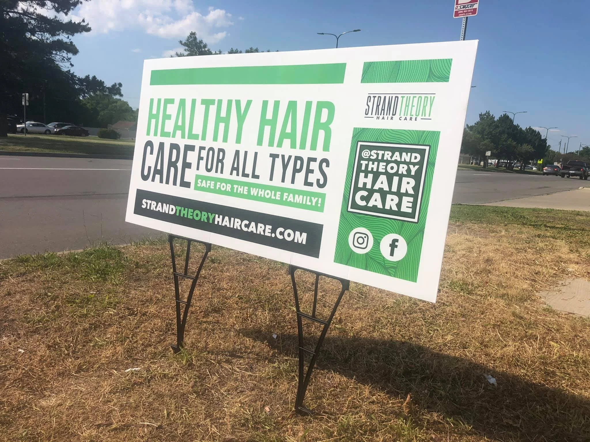A sign on the grassy area beside a road advertising Strand Theory Hair Care, emphasizing healthy hair care for all types and the whole family, with social media icons for Instagram and Facebook.
