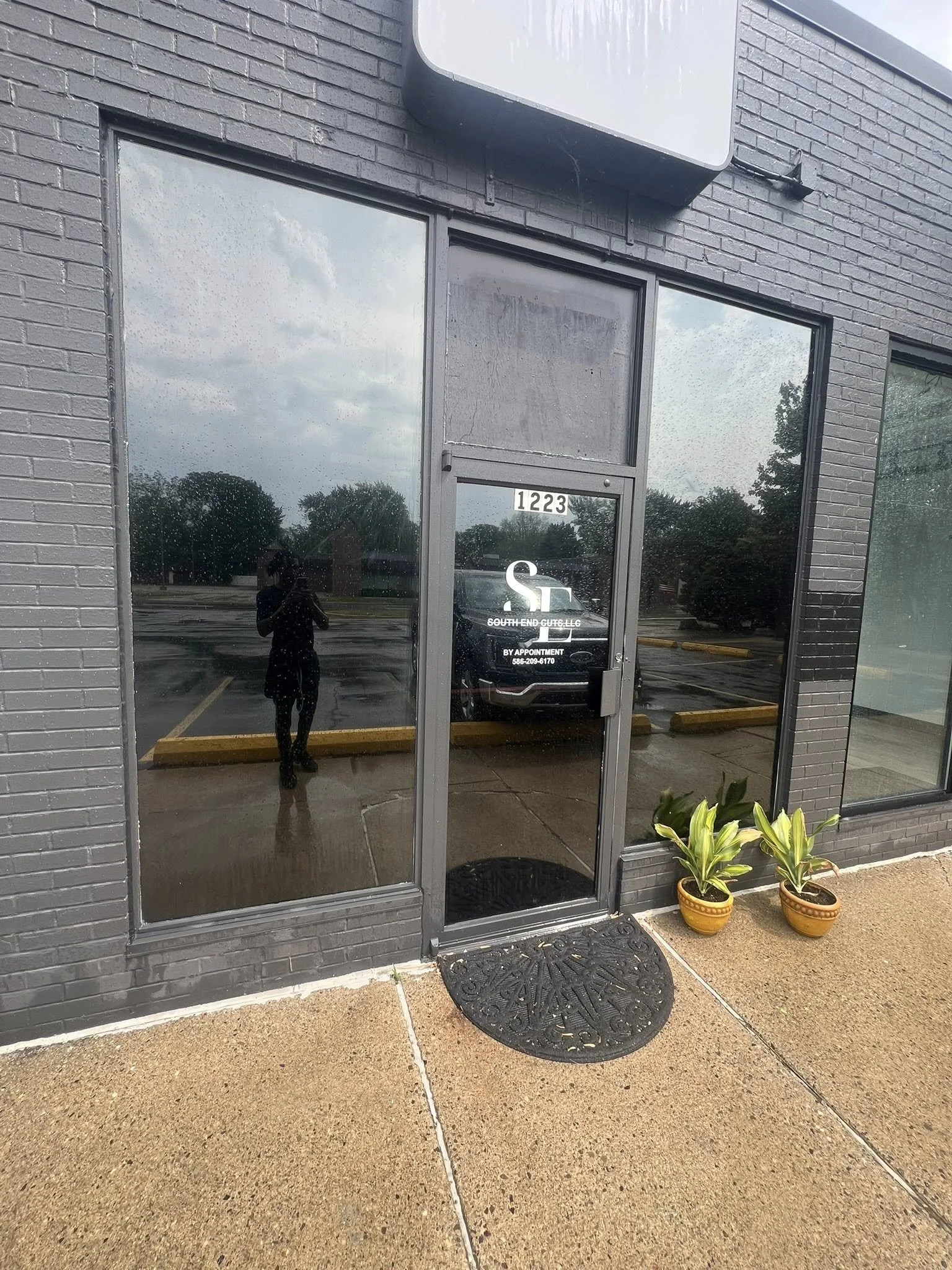 Storefront with black brick exterior, glass door with white signage, and two potted plants outside, with a parking lot and cloudy sky reflected in the glass.