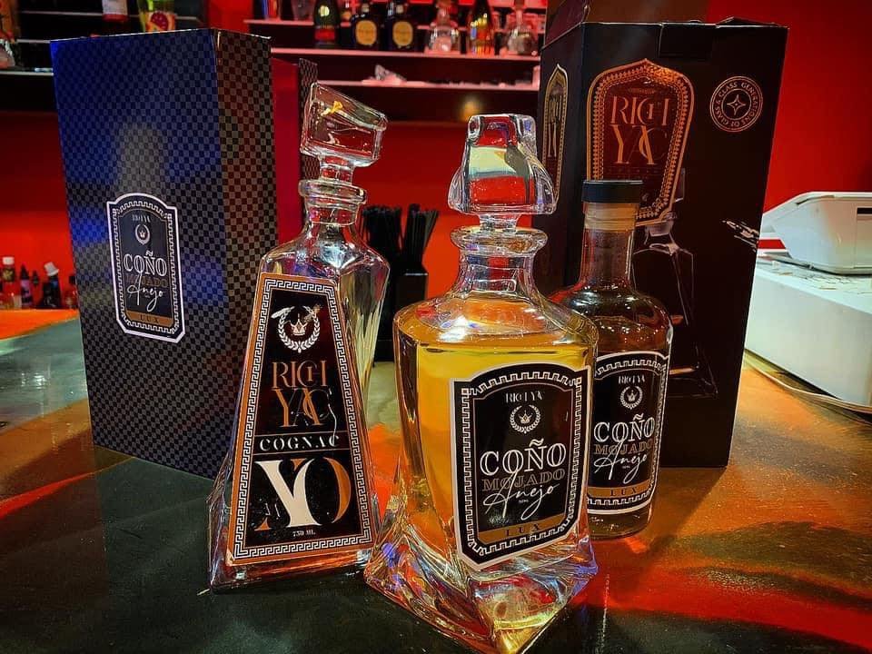 Three bottles of cognac with black labels and gold and white text, positioned in front of decorative boxes on a bar counter.