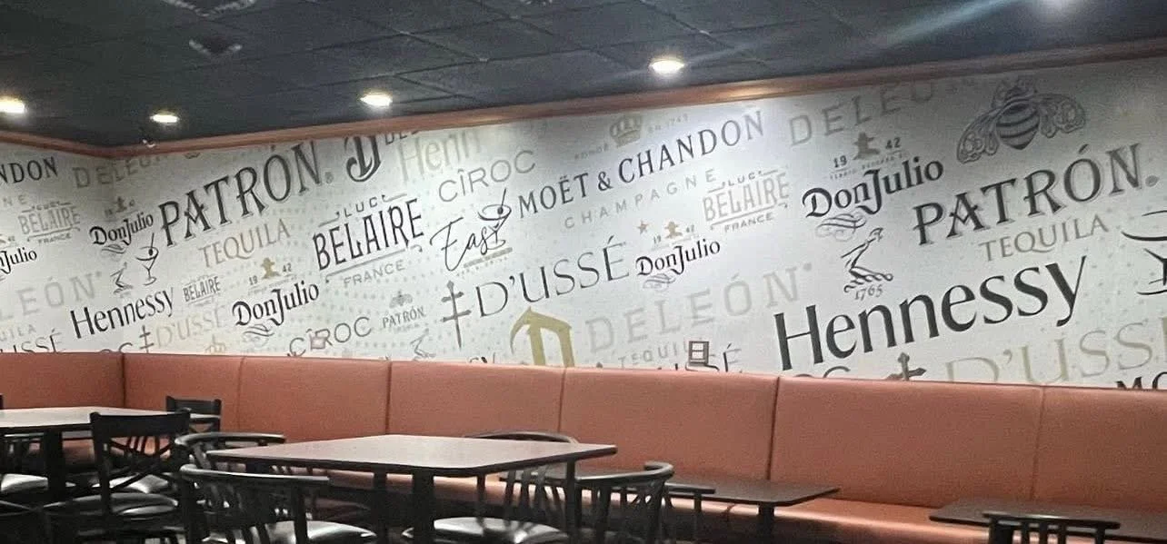 Indoor restaurant with a high cushioned pink bench along a white wall decorated with various alcohol brand logos and text.