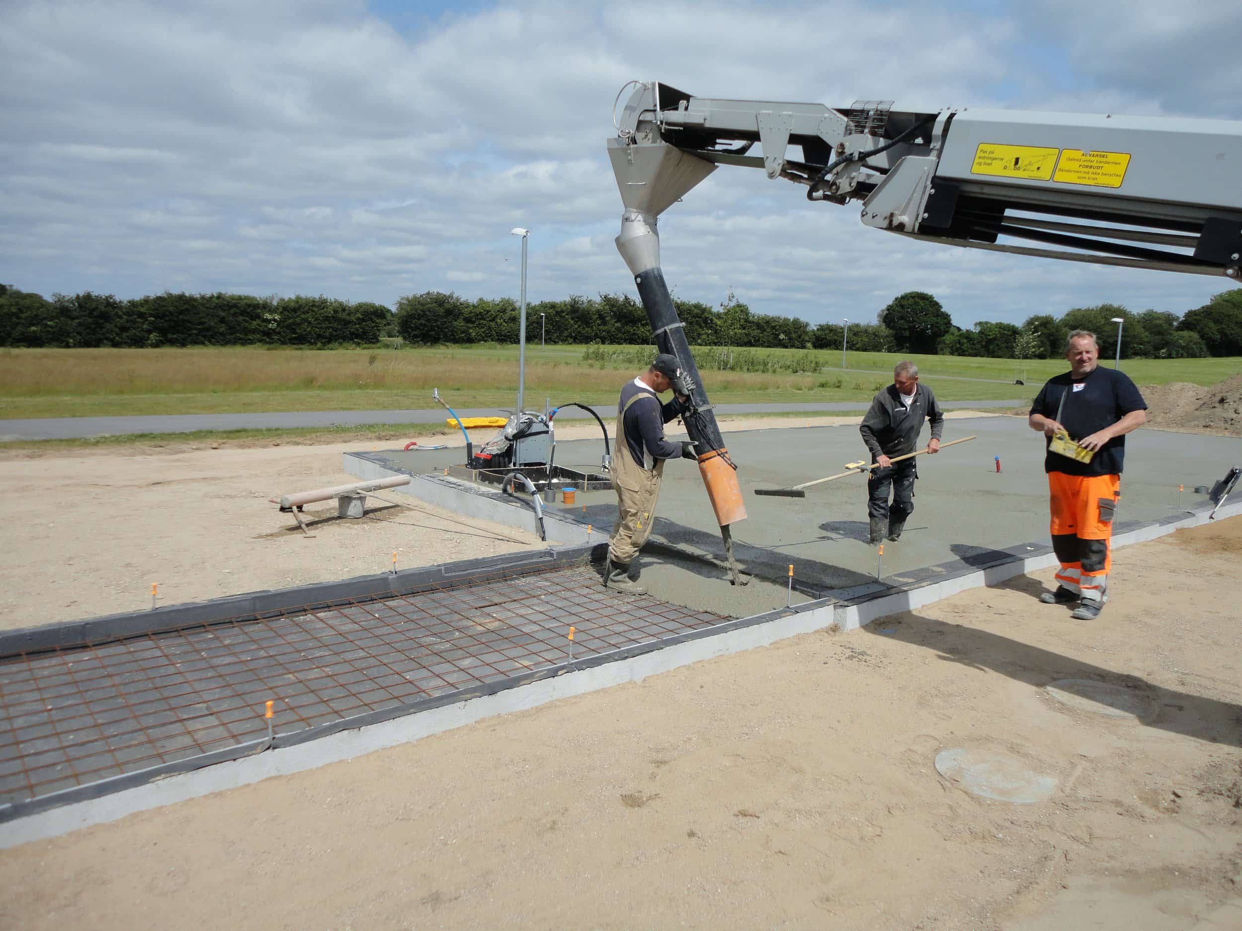 Folk arbejder på en cementflade, der bliver støbt, mens en kran hælder beton over området. Der er tre mænd i arbejdstøj, og de bruger værktøj til at glatte betonen ud.