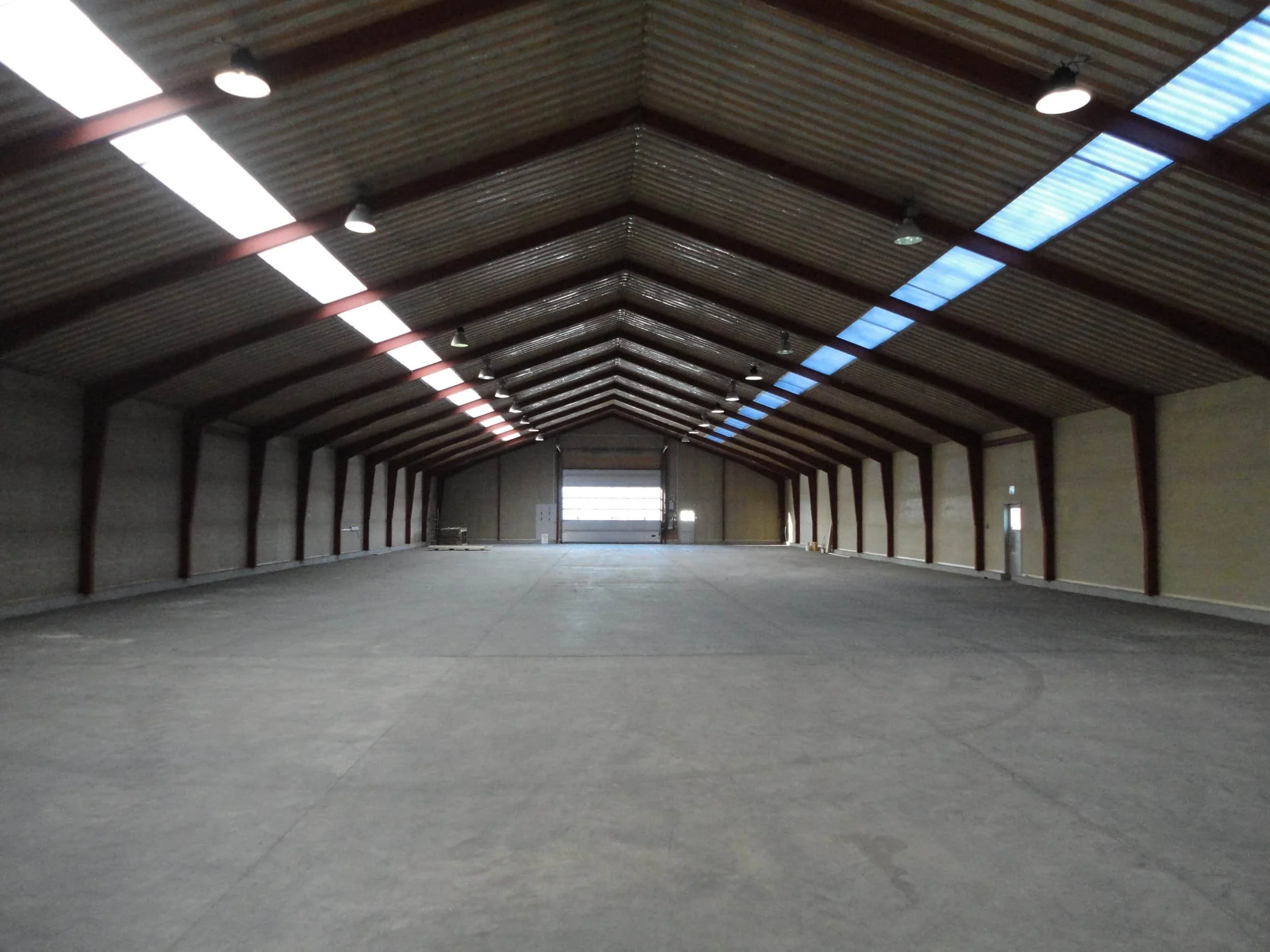 Et stort tomt industrilager med betonbund, høj buet metalstruktur, træpanel på væggene, og belysning i loftet, indgangen i enden.