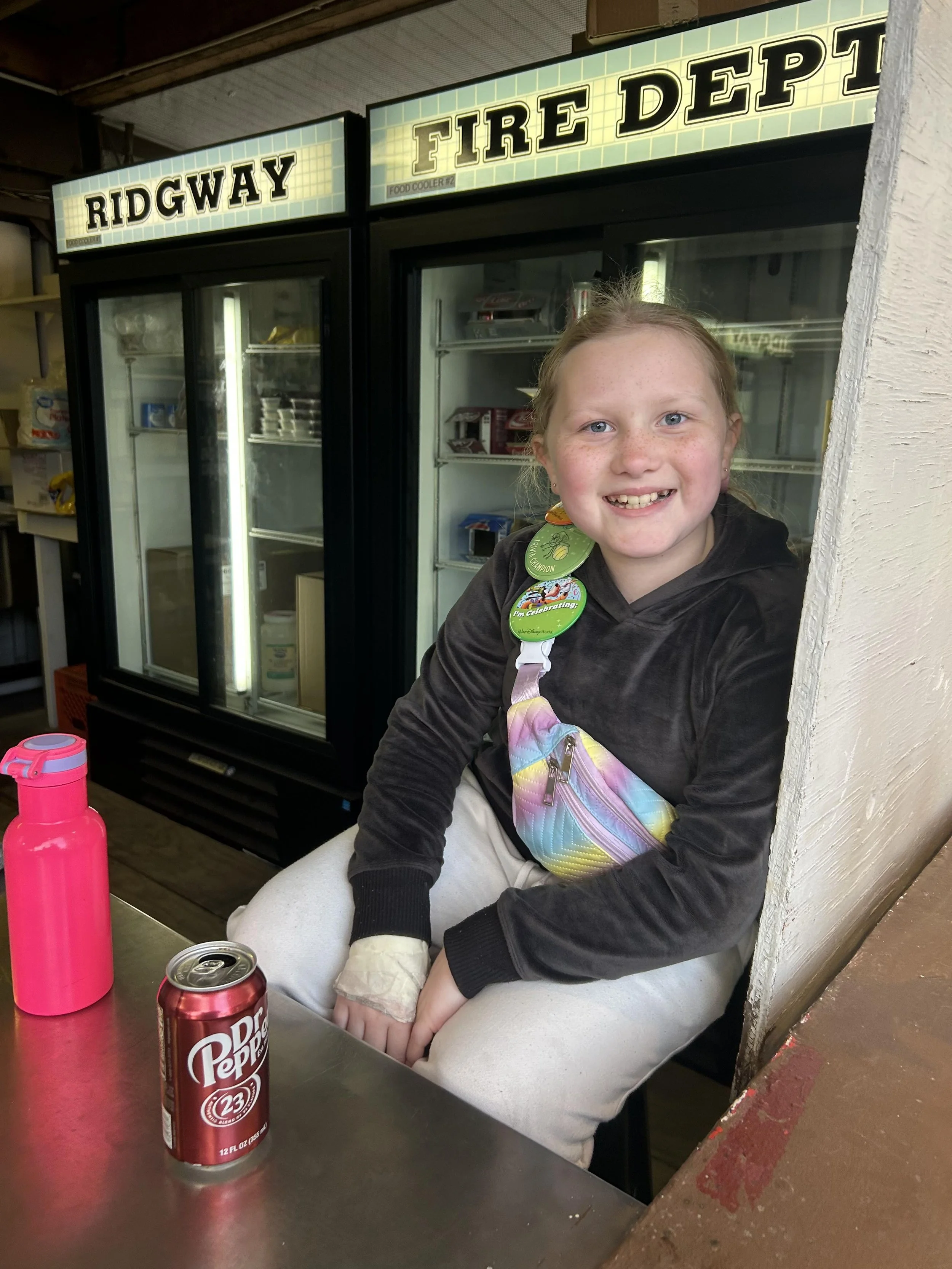A young girl with curly red hair and freckles, smiling, sits at a table with a pink water bottle and a can of Dr. Pepper in front of her. She is wearing a black hoodie, white pants, and colorful accessories around her neck. Behind her are refrigerators labeled 'RIDGWAY' and 'FIRE DEPT.' in a casual setting.