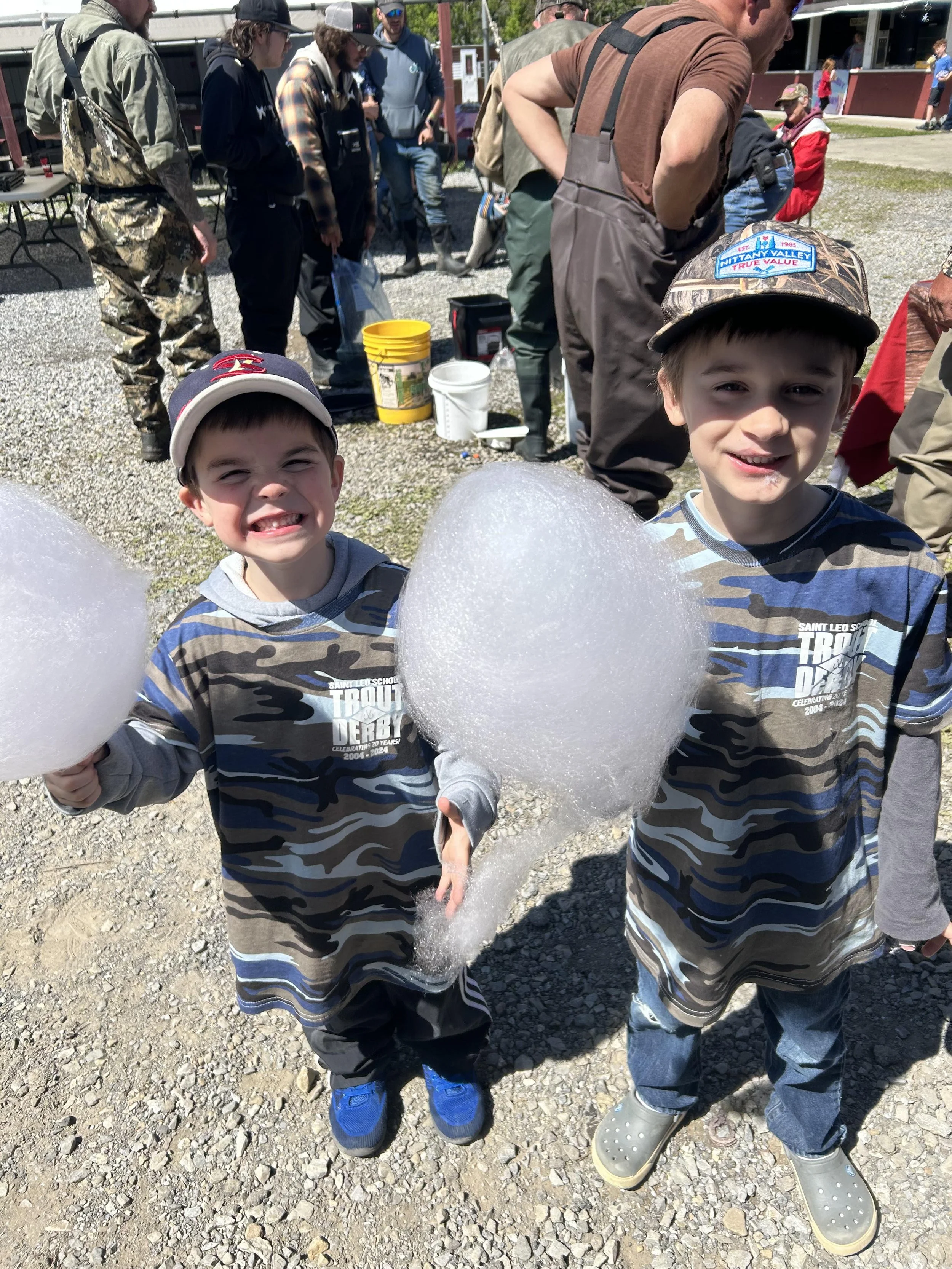 Two young boys smiling and holding large cotton candy sticks at an outdoor event on a sunny day, with other people in the background.