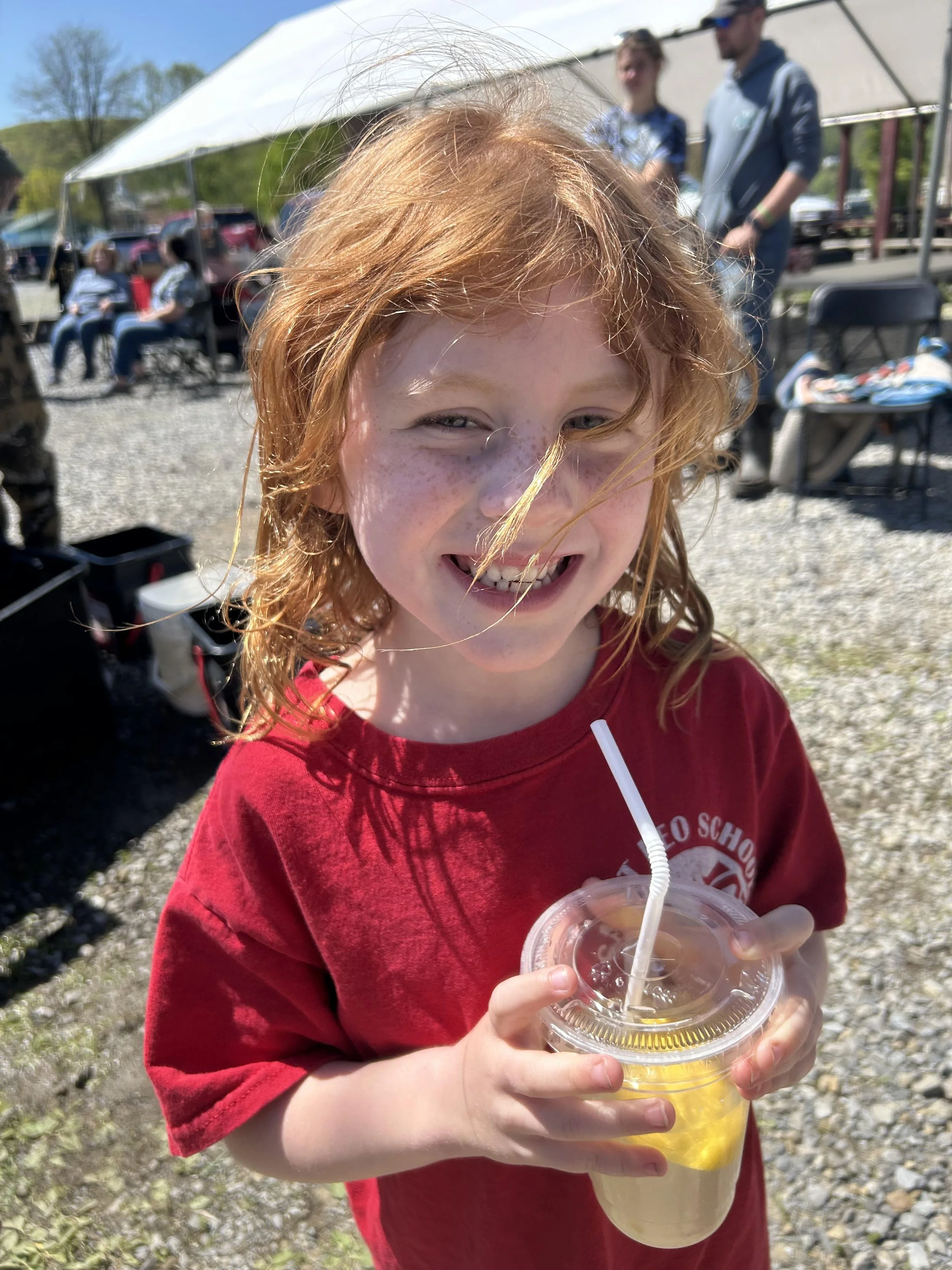 Smiling red-haired girl holding a cup with a straw outdoors at a sunny event, with people and a tent in the background.