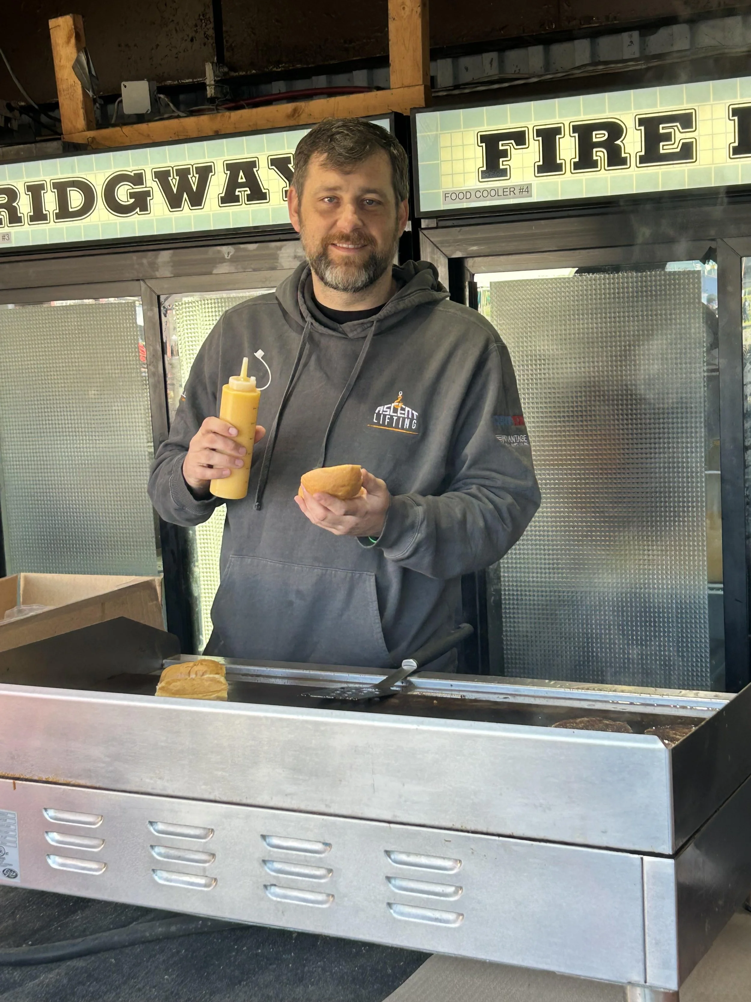Man standing behind a grill holding a hot dog and mustard bottle, with signs reading 'RIDGWAY' and 'FIRE' in the background.