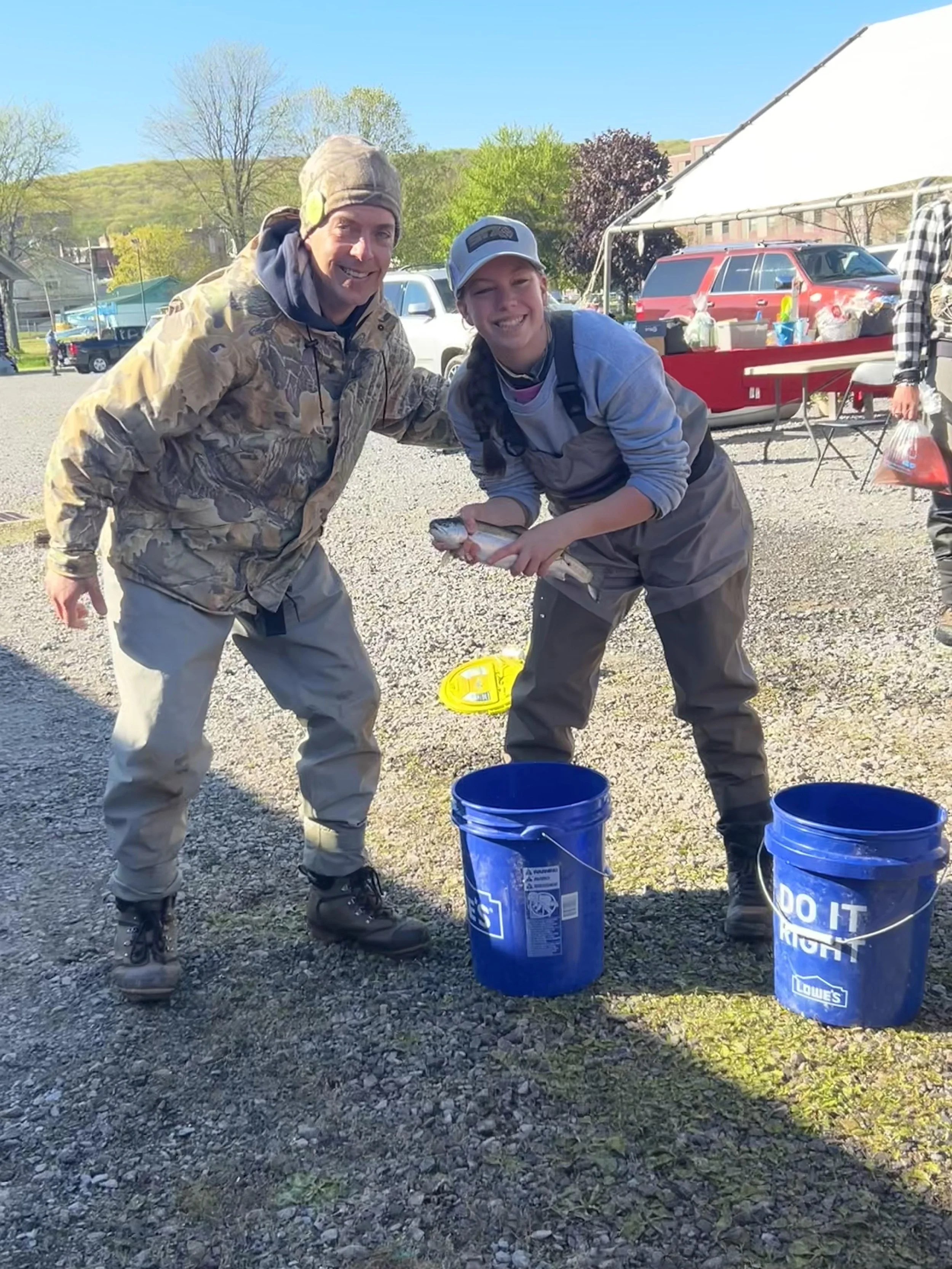 Two men, one older and one younger, are outdoors smiling and holding a fish between them. They are dressed in outdoor clothing and boots. There are two blue buckets on the ground nearby, and there are cars and a tent with tables in the background.