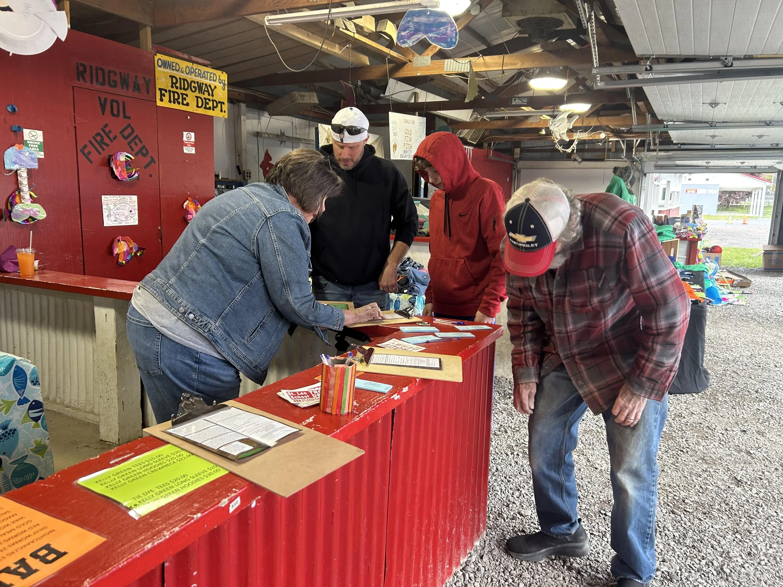 Four people are at a red counter inside a building, with one woman and three men. The woman is writing on a paper while the others look on. The background shows a red wall with signs and decorations, and the exterior view through an open garage door.