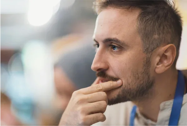 A man with short brown hair and a beard, wearing a white shirt with blue accents, is resting his chin on his hand and appears to be thinking.