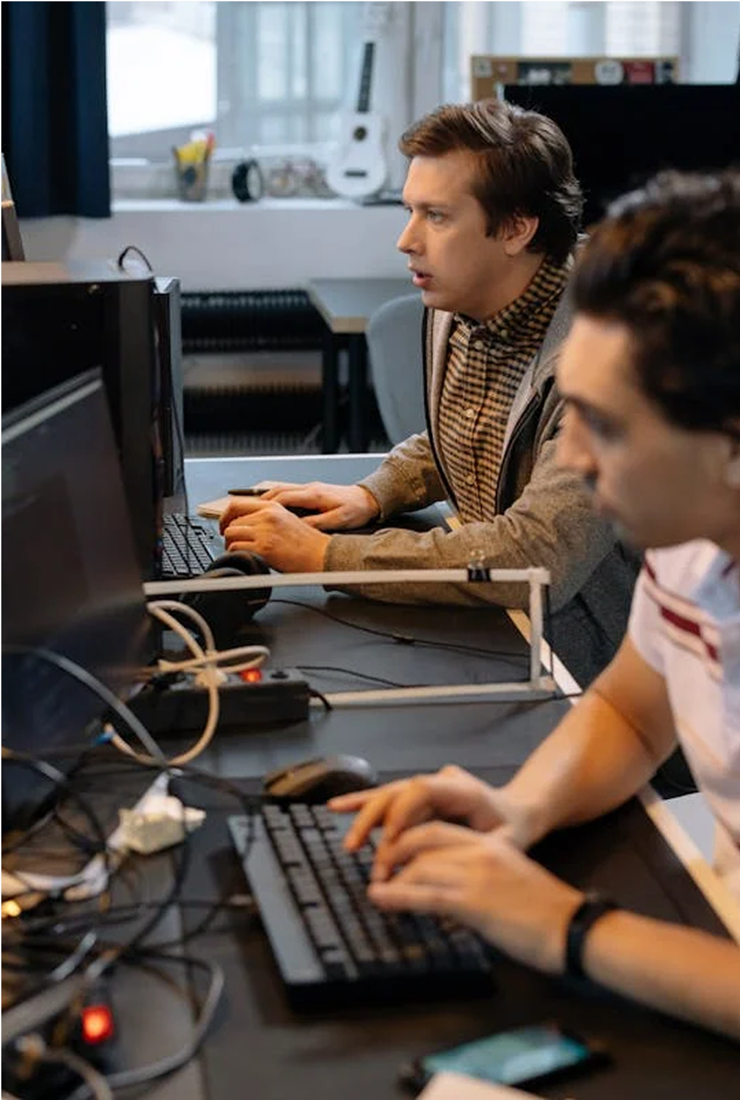 Two men working at computer stations in an office, with various wires and electronic devices on the desk.