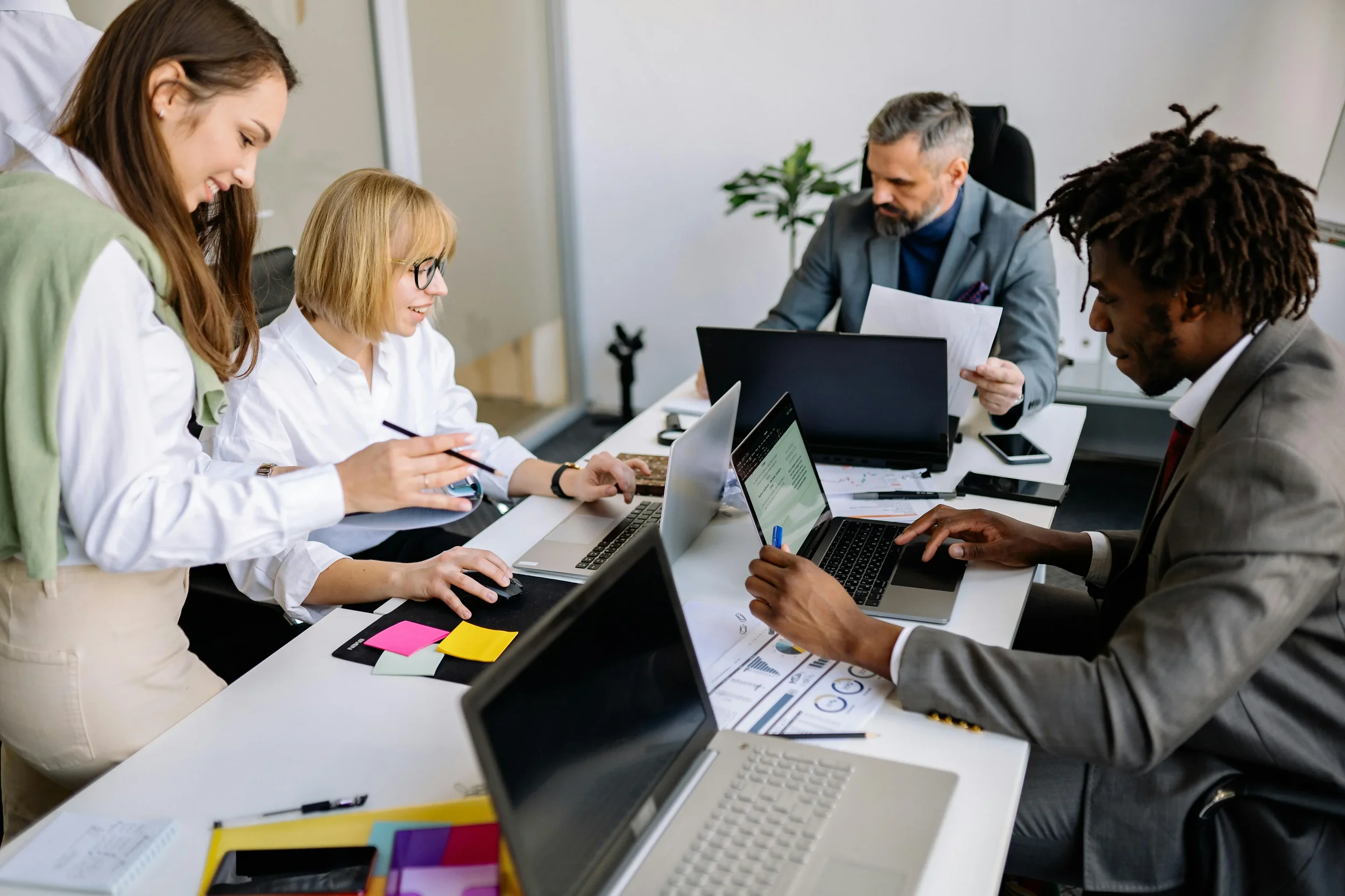 A diverse group of five professionals in a meeting, working on laptops and discussing, with documents, notebooks, and office supplies on the table.