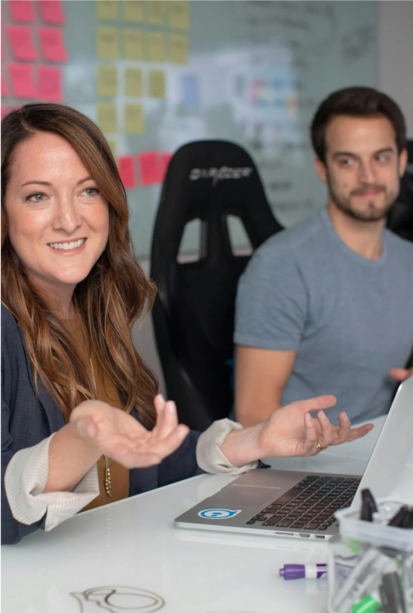 A woman with long brown hair smiling and gesturing with her hands while sitting at a table with a laptop in front of her. A man with short dark hair and a beard, wearing a gray T-shirt, is sitting beside her looking slightly to the side. In the background, there is a whiteboard covered with colorful sticky notes.