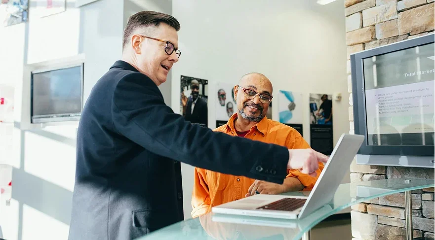 Two men smiling and talking in an office or conference room, one pointing at a laptop screen, with monitors on the wall behind them.