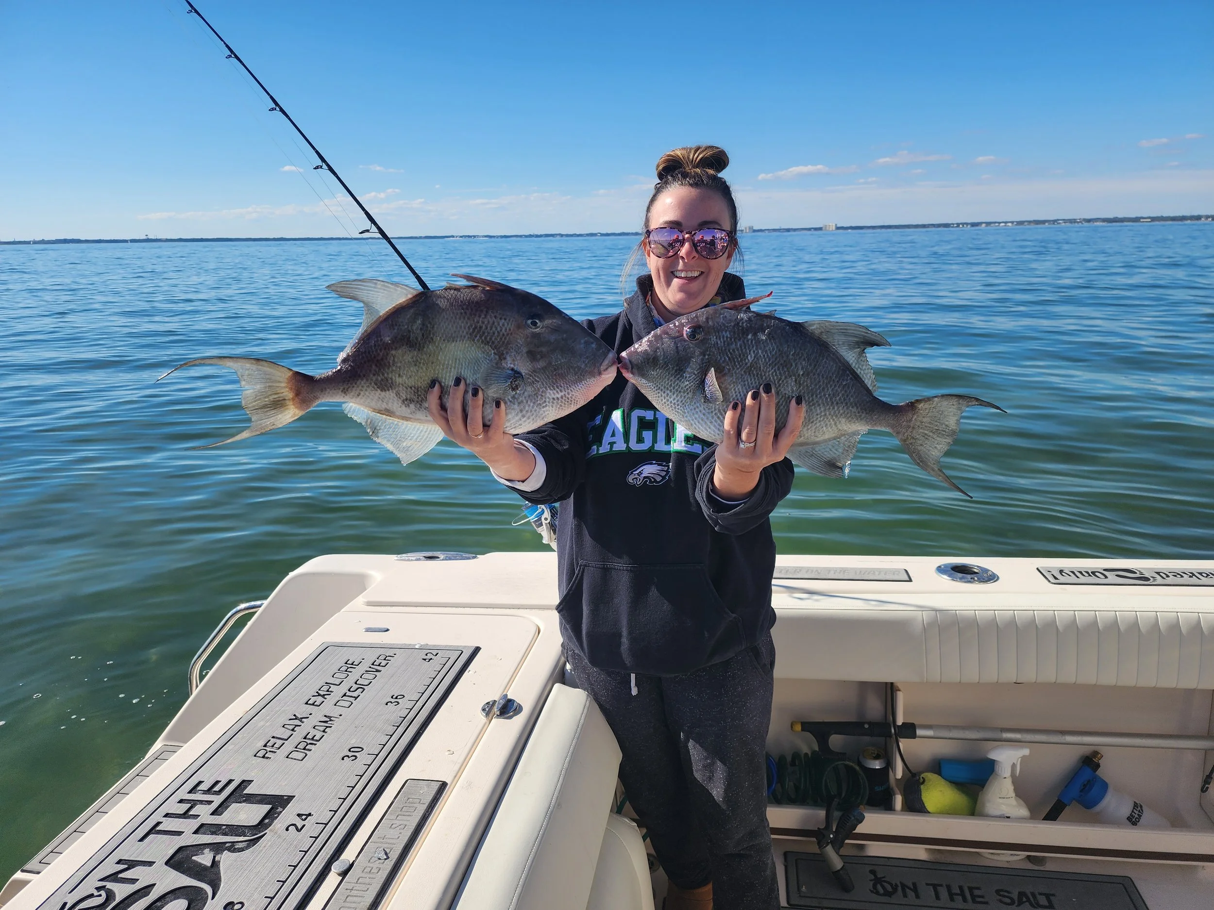Woman on a boat holding two large fish with a body of water and blue sky in the background.