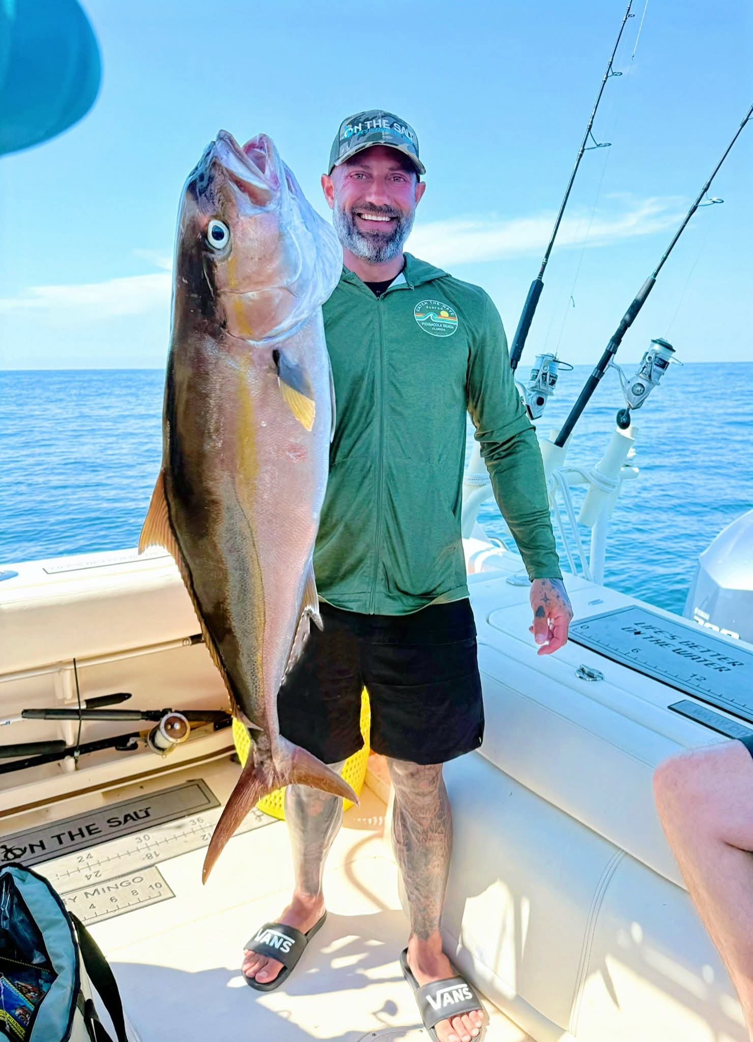 A man smiling and holding a large fish on a boat in the ocean, with fishing rods in the background.