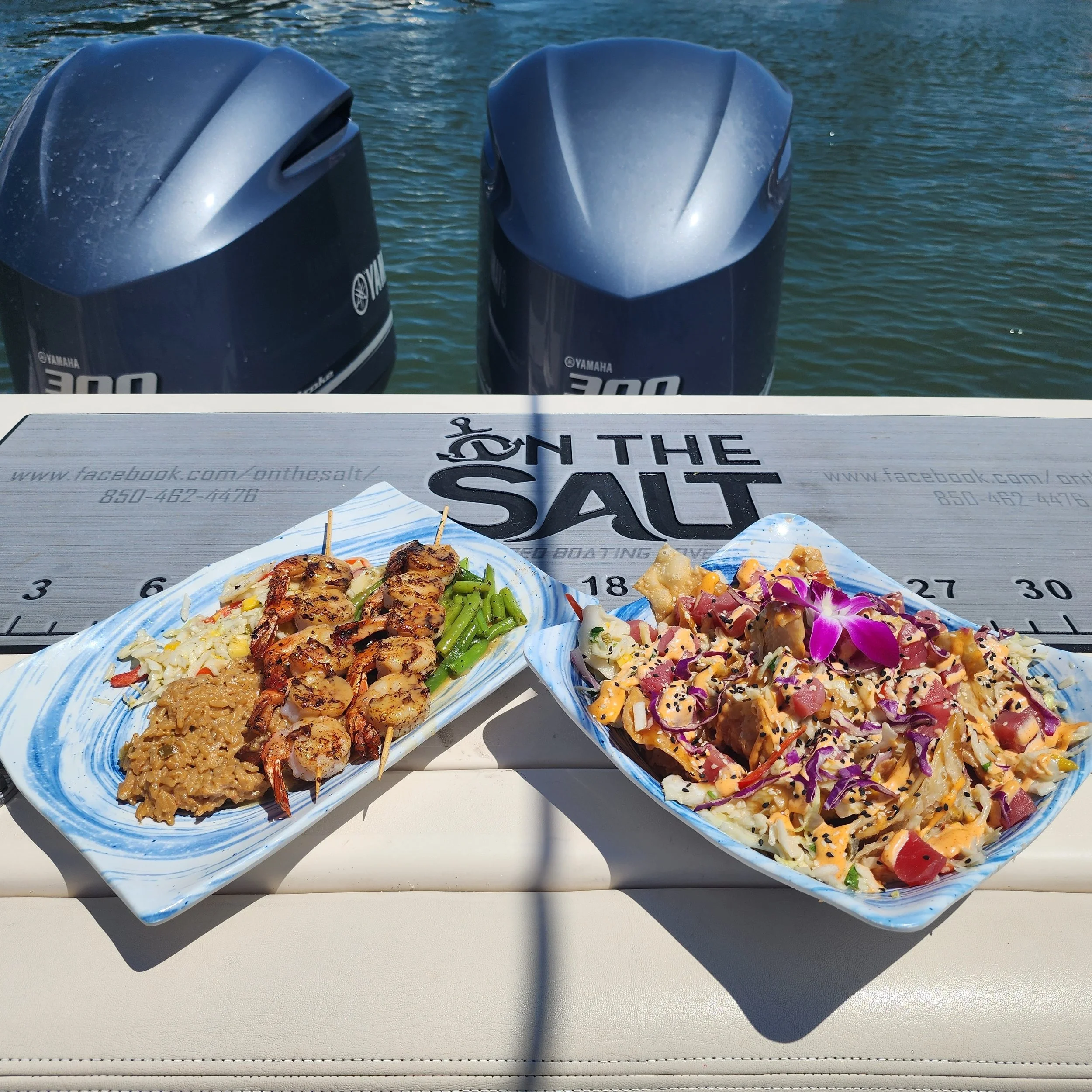 Two plates of food on a boat with a water background and two Yamaha outboard motors. The plates contain skewers of grilled meat, rice, and vegetables, and a colorful mixed salad with purple flowers.