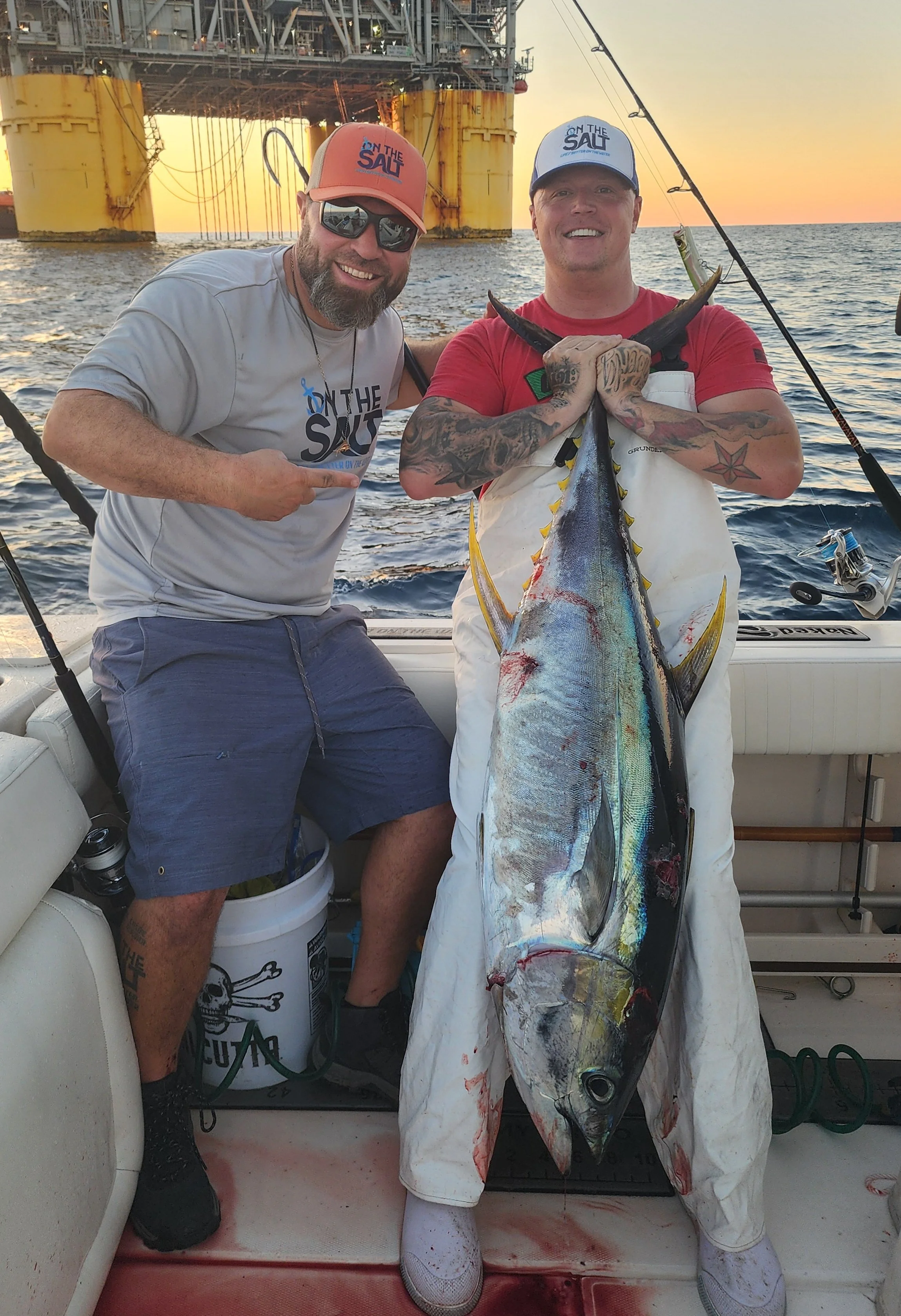 Two men on a boat holding a large yellowfin tuna, with an offshore oil rig in the background during sunset.