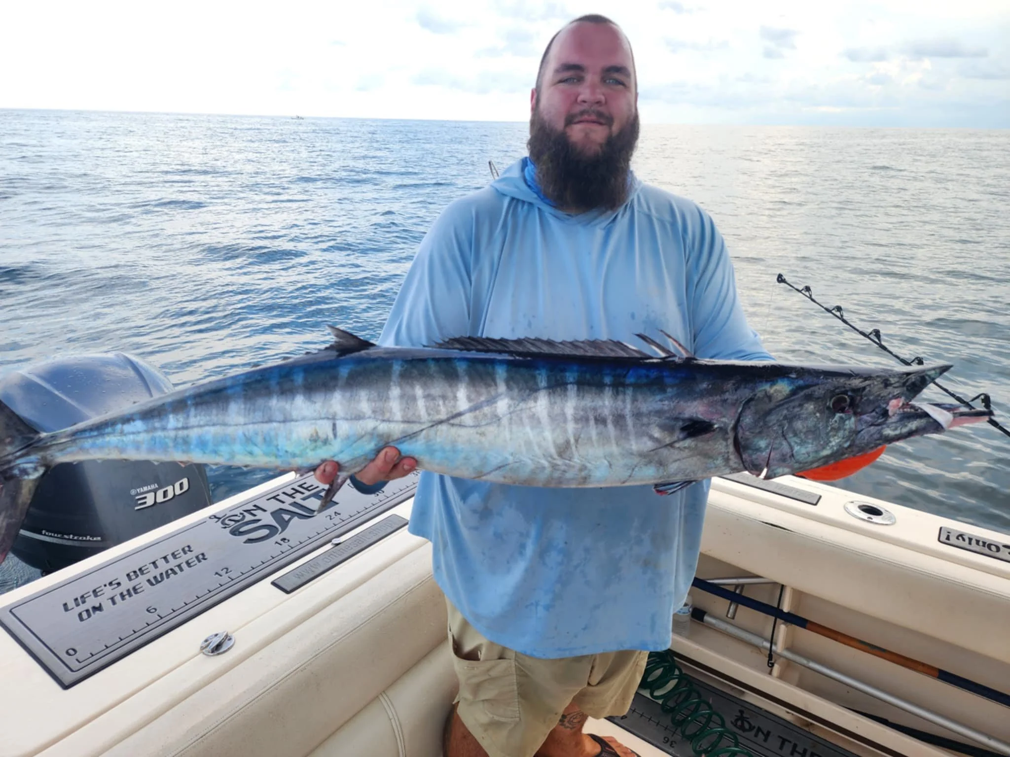 Man with a beard in a light blue shirt and khaki shorts on a boat, holding a large fish with a long, streamlined body and blue and silver stripes, over a calm ocean under a cloudy sky.