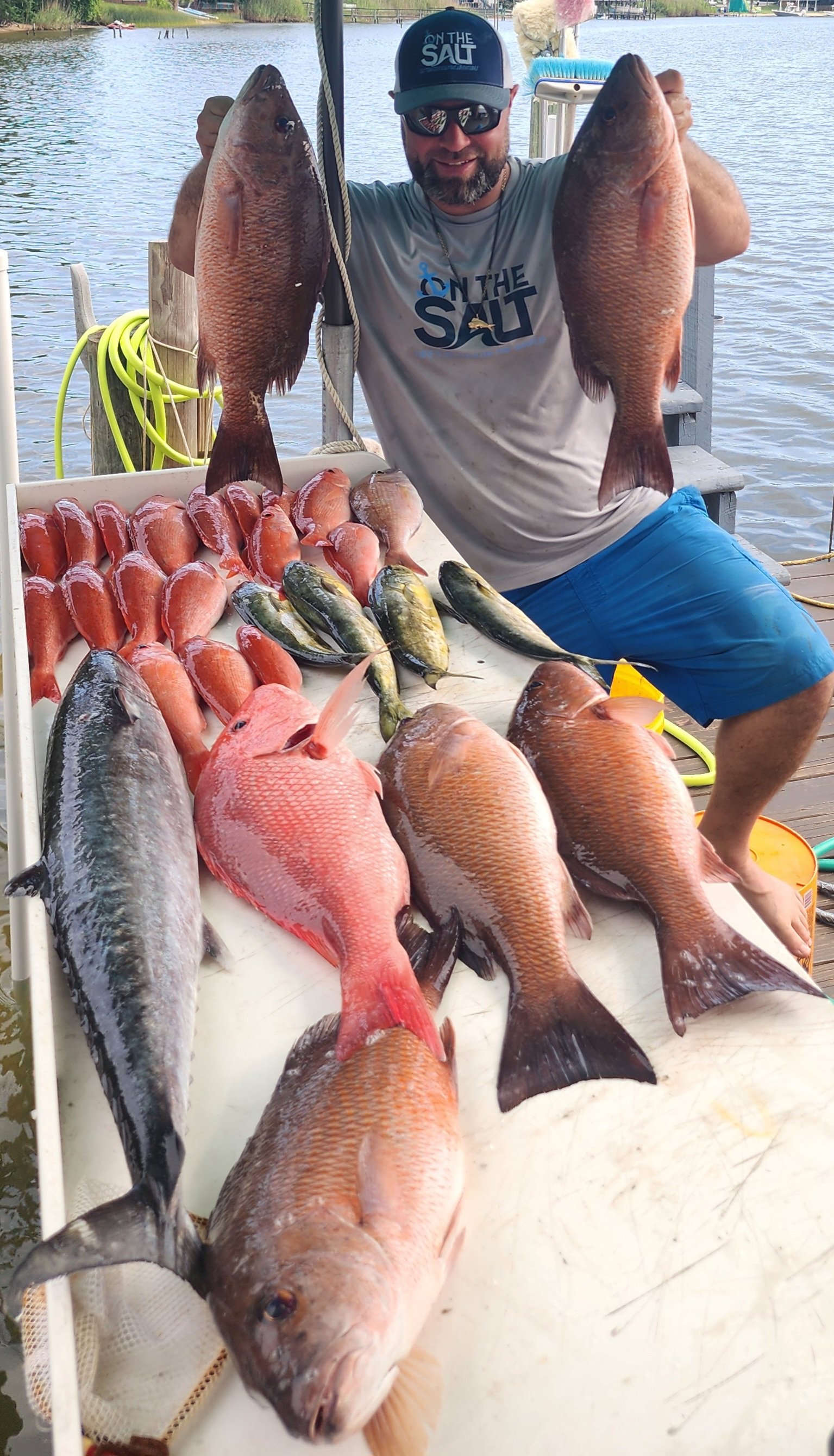 Man holding two large fish, with a table of various smaller fish and other catch, on a dock near a body of water.