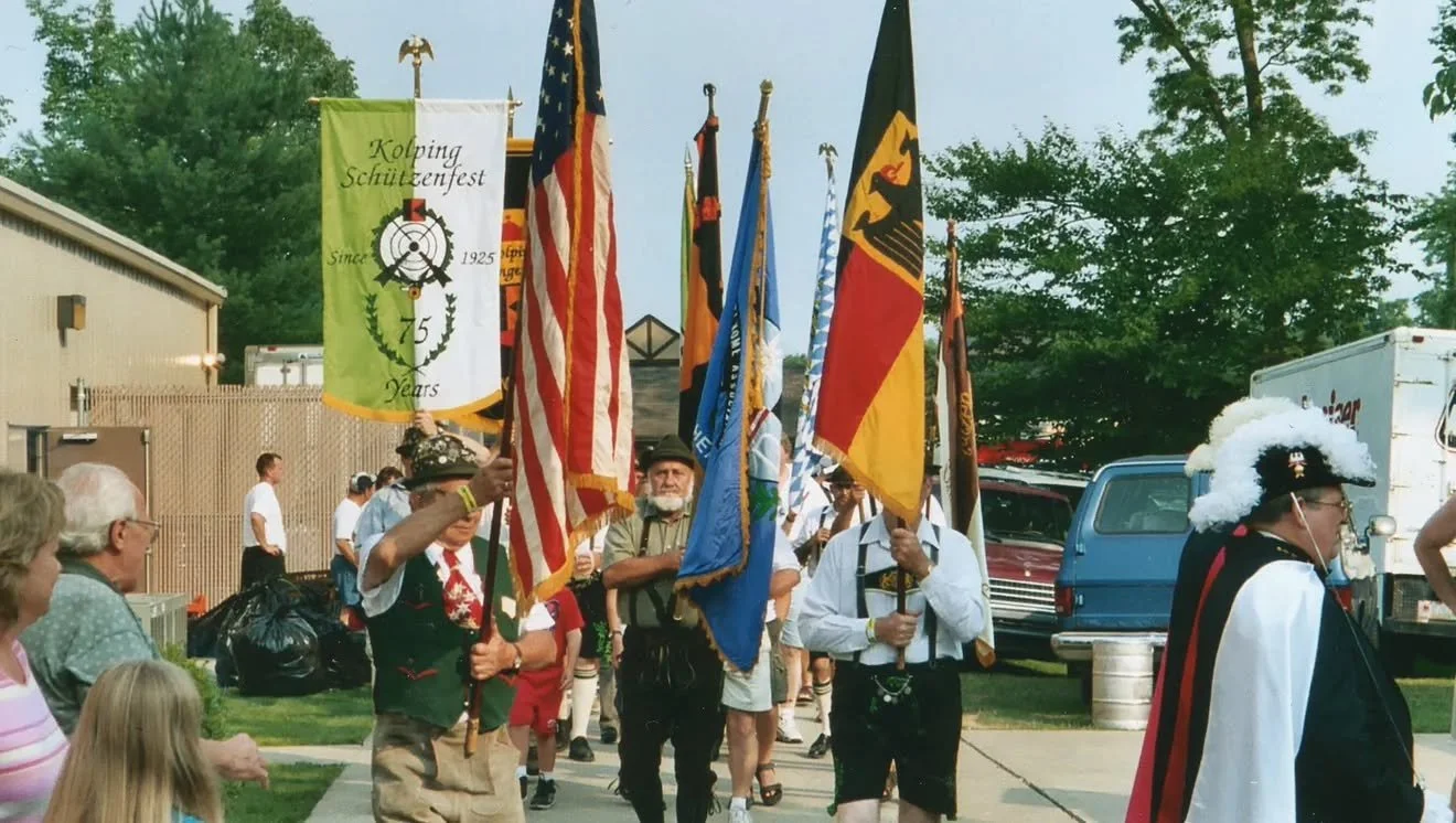 Group of people participating in a parade or event, carrying various flags including American and German flags, with onlookers watching.