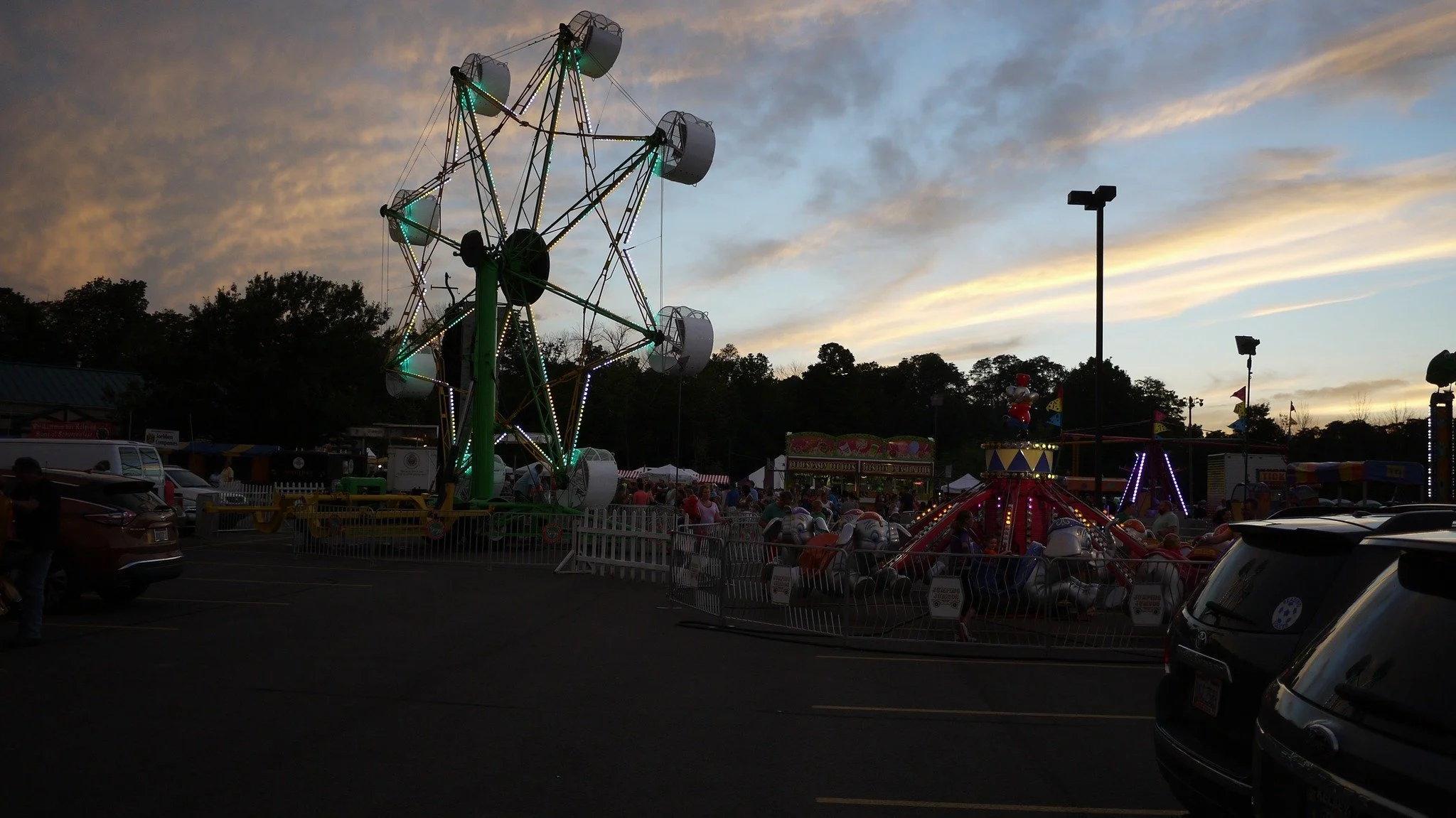 A carnival at dusk with amusement rides, including a Ferris wheel with lights, and a carousel with children. There are parked cars in the foreground, and the sky is partly cloudy with the sunset.