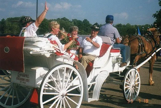 Group of people riding in a white horse-drawn carriage, smiling and waving as they travel on a sunny day.