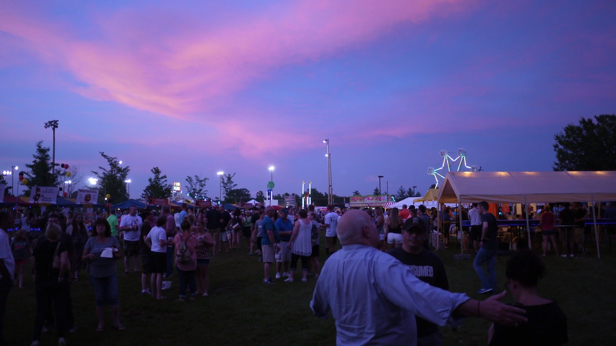 Crowd of people at a fairground during dusk with colorful sky, tents, and amusement rides in the background.