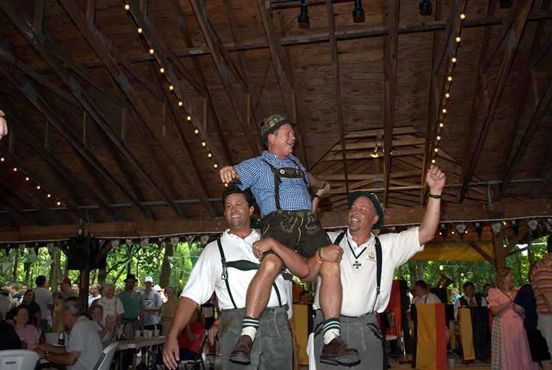 People celebrating inside a wooden hall decorated with string lights, with a man sitting on the shoulders of two others, raising his fist in the air, wearing traditional German attire.