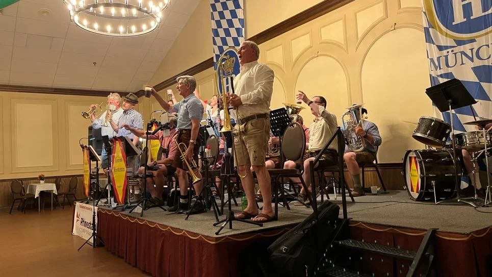A band of musicians performing on a stage decorated with Bavarian flags and banners, with some members playing brass instruments and drums, and one person standing with a large beer mug.