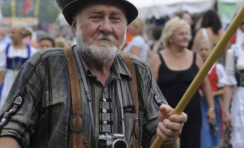 An elderly man with a gray beard, wearing a hat and plaid shirt, holding a long pole at an outdoor event with several women in the background.