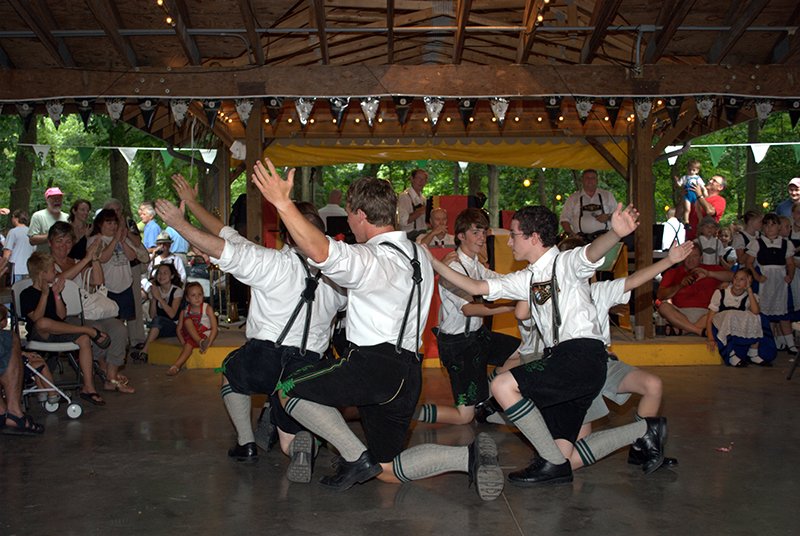 Group of young men performing a traditional dance in a wooden pavilion, with audience watching and some dressed in folk costumes.