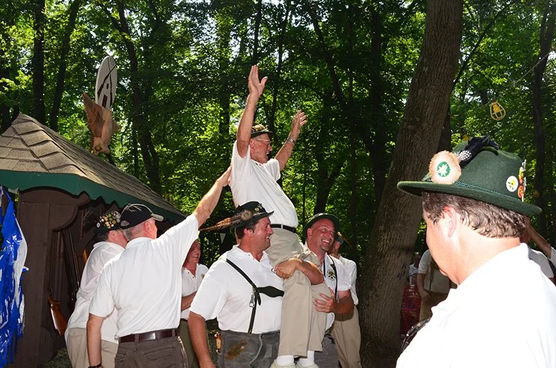 A group of men dressed in traditional Bavarian clothing celebrating outdoors with trees in the background, some wearing hats and lederhosen, holding a man up in the air.