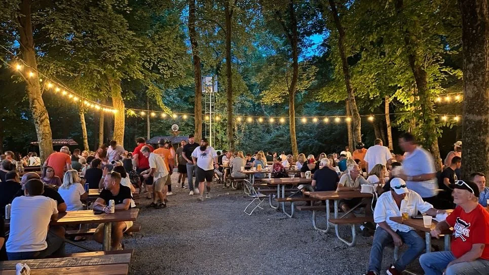 An outdoor gathering at dusk with many people sitting at wooden picnic tables under string lights in a wooded area.