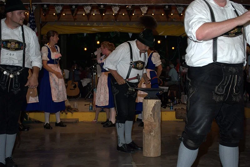 Group of people dressed in traditional German folk costumes, dancing and playing music at an outdoor event under string lights, with a man chopping wood during a performance.