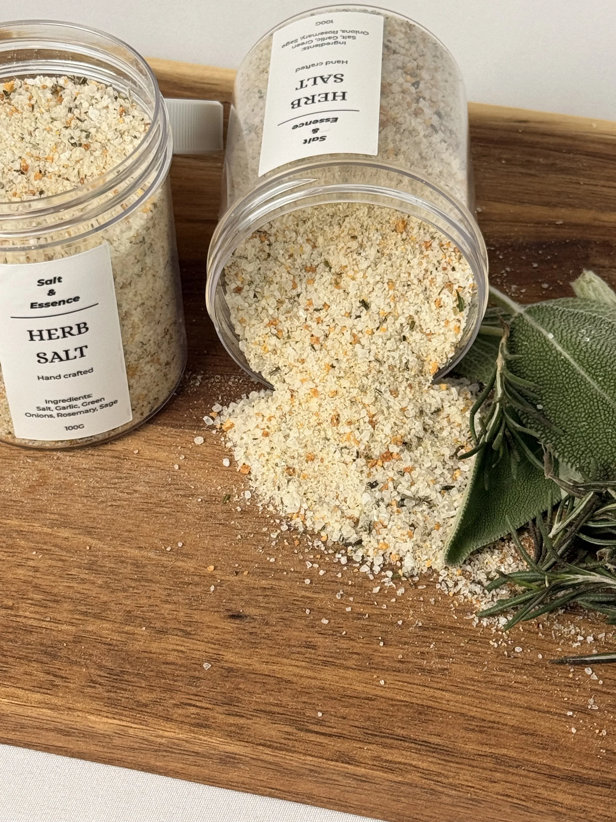 Various jars of herb salt, some spilled on a wooden surface, with sprigs of fresh herbs nearby.