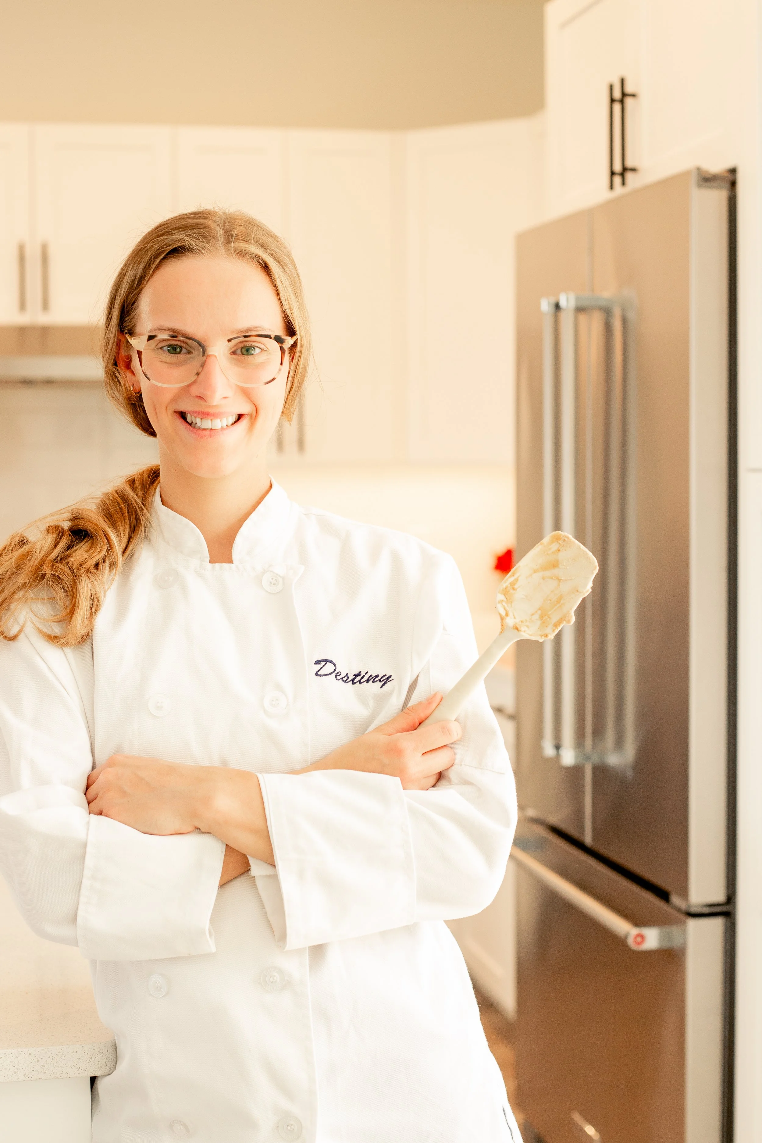 A woman wearing a white chef's coat with 'Destiny' embroidered on it, smiling, holding a spatula, standing in a modern kitchen with white cabinets and a stainless steel refrigerator.
