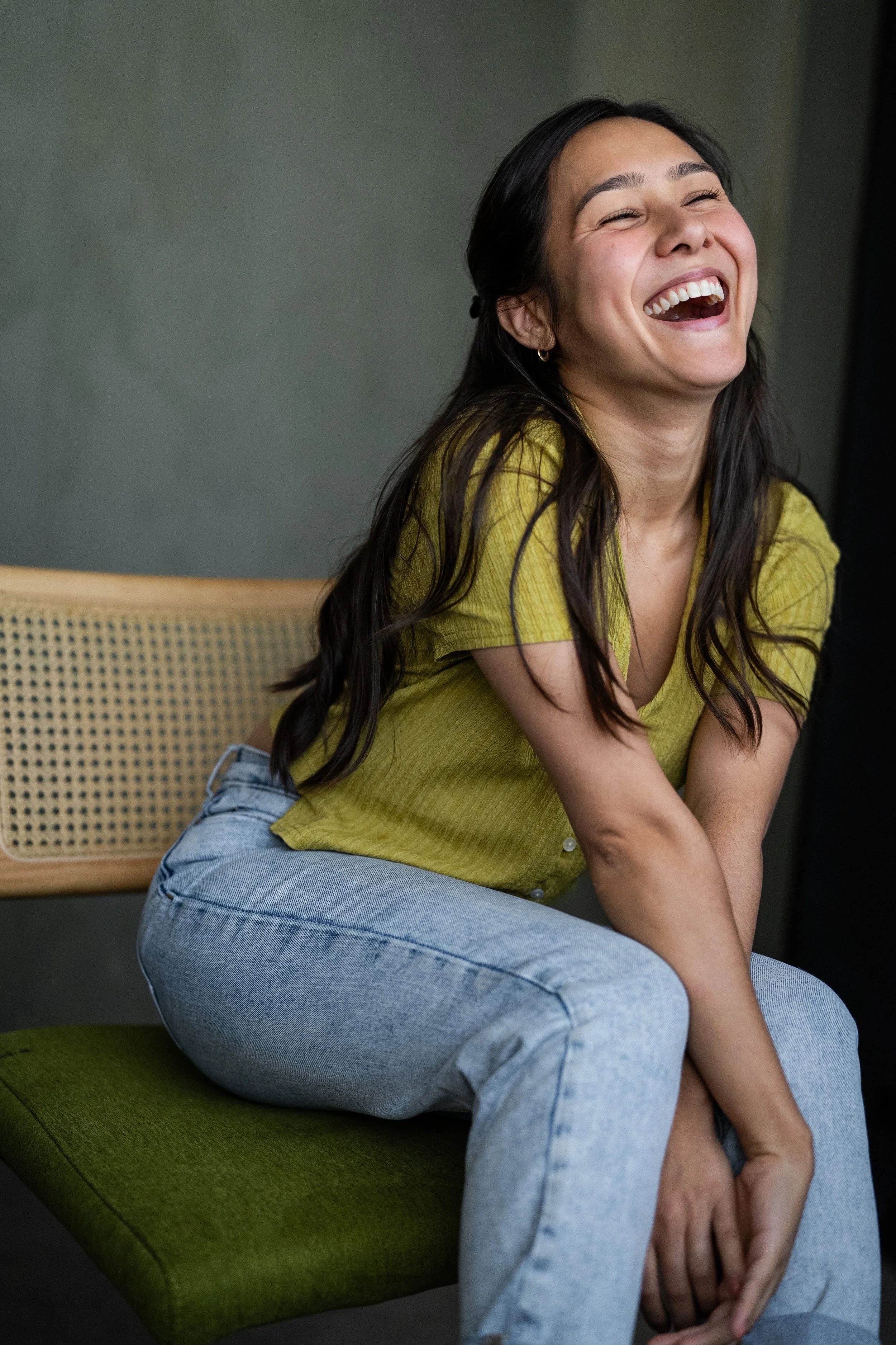 Actor Marielle Young sitting on a green cushioned chair, laughing with her head tilted back and eyes closed, wearing a yellow shirt and light blue jeans.