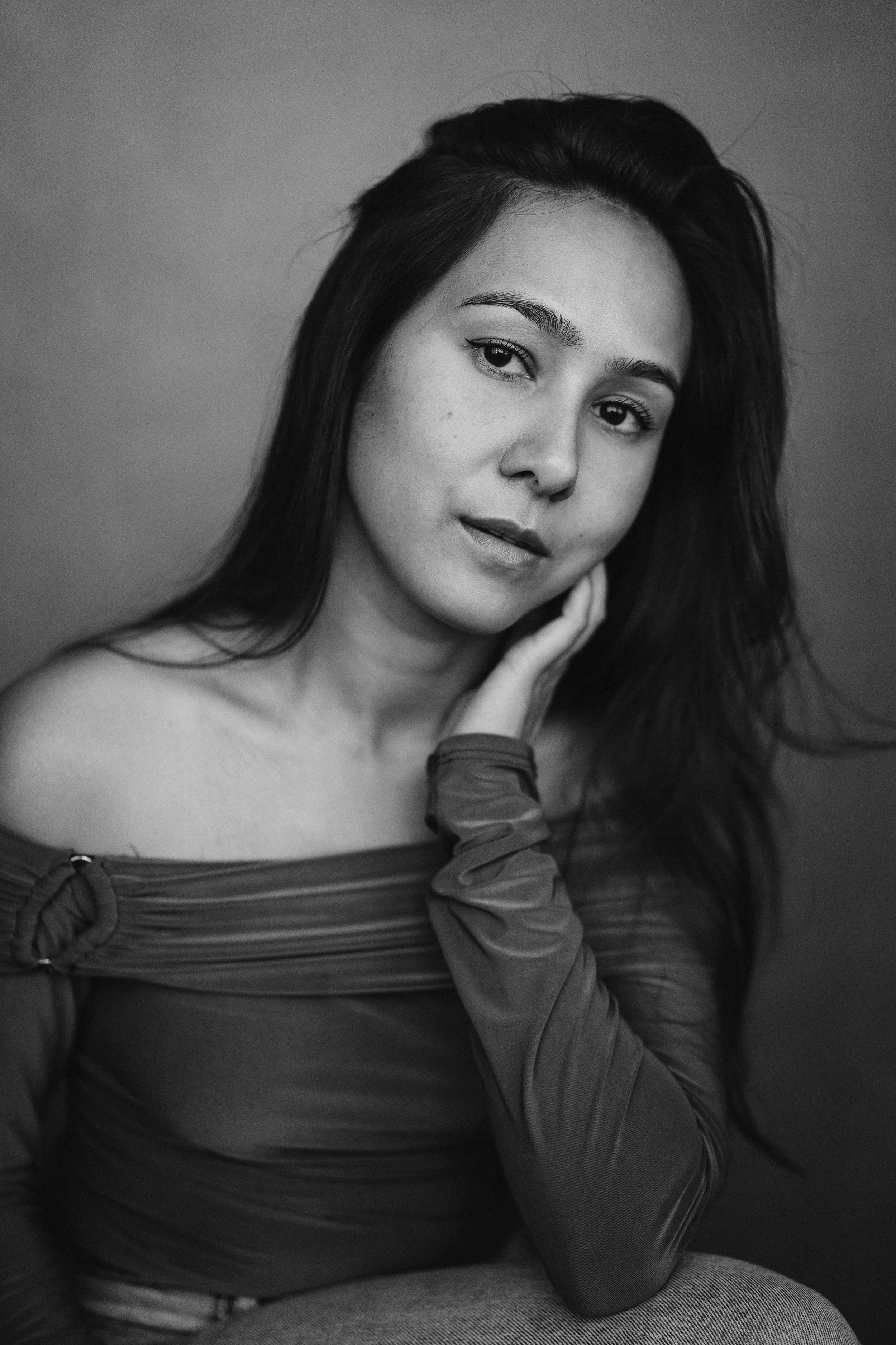 Black and white portrait of actor Marielle Young with long dark hair, wearing an off-the-shoulder top, looking at the camera with a relaxed expression, touching her face with her left hand.