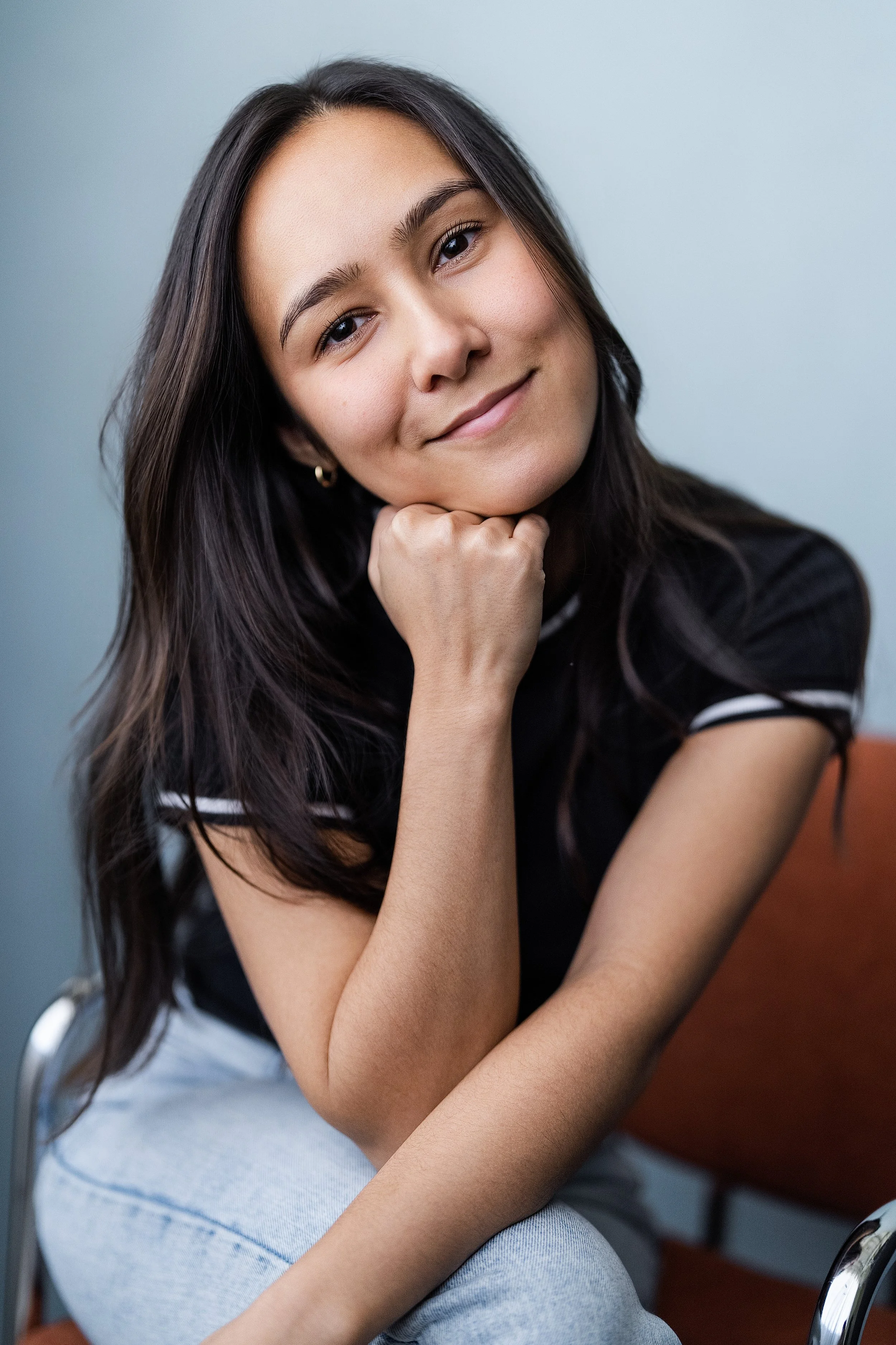 Actor Marielle Young with long dark hair smiling and resting her chin on her hand, sitting on a chair.