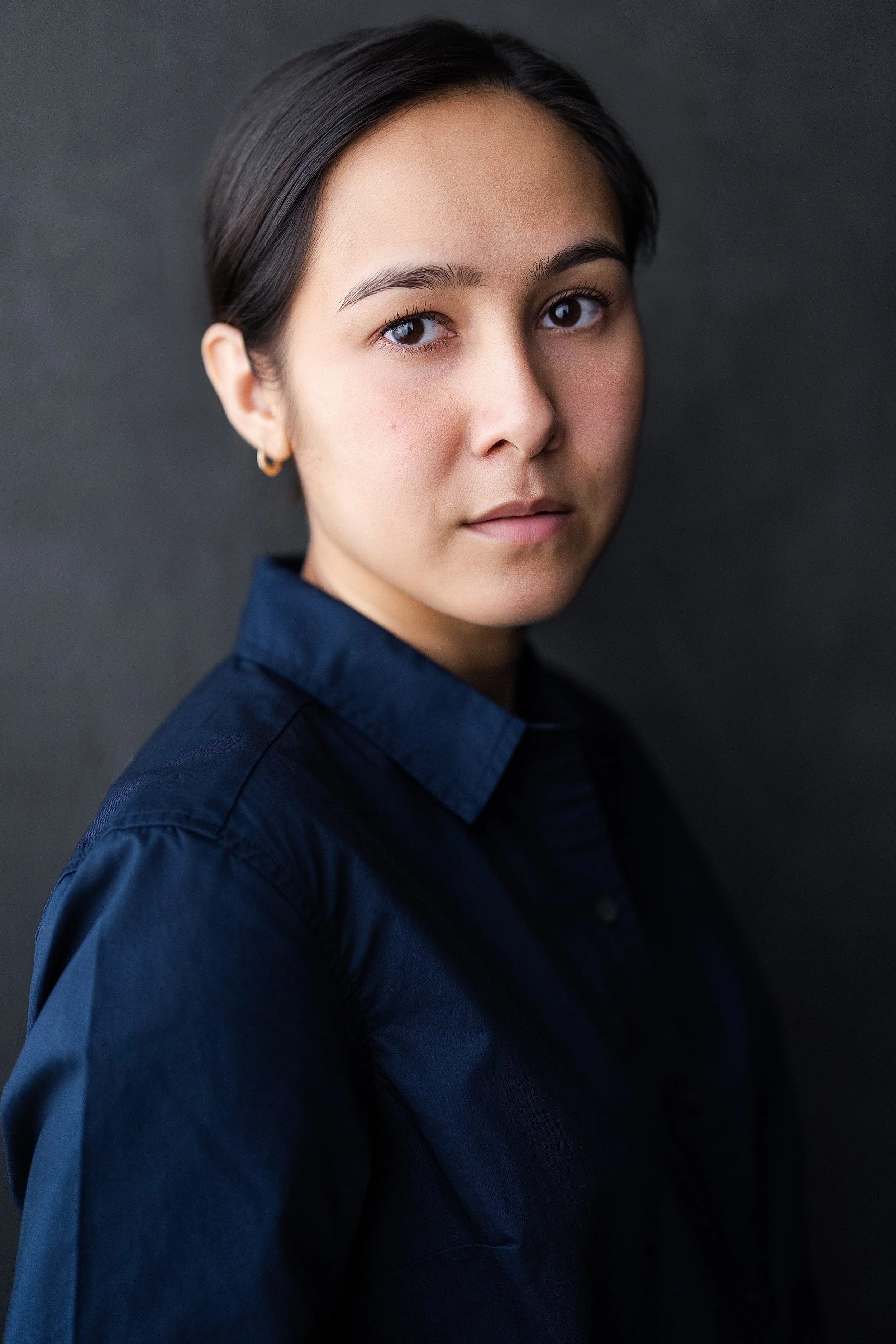 Portrait of actor Marielle Young with dark hair, wearing a dark blue shirt, standing against a dark background.